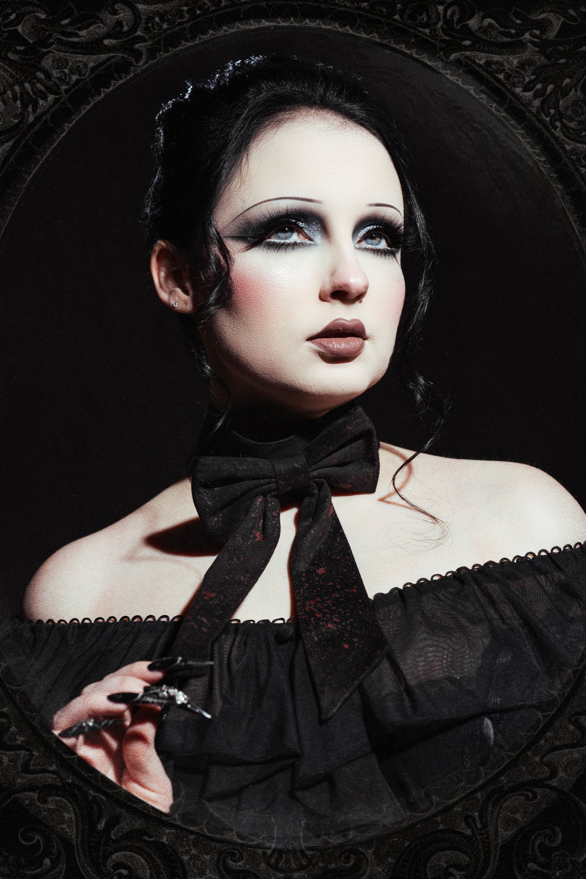 A gothic-style portrait of a woman with pale makeup, dramatic dark eye makeup, and a black off-the-shoulder dress with a large black and red bow choker necklace, framed by an ornate dark oval border.