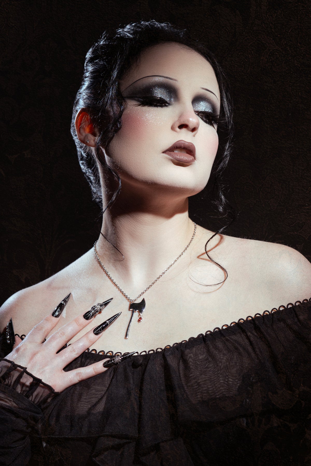 woman with dark eye makeup wearing a black off the shoulder dress with a silver necklace that has an axe pendant and red gem