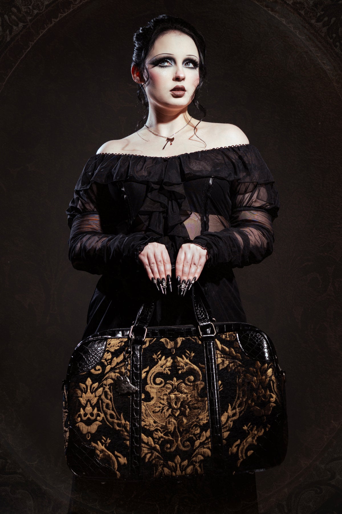 A gothic-style woman with pale makeup and dark hair models a black off-the-shoulder sheer long-sleeve dress while holding a large black and gold damask-patterned duffel bag against a dark, ornate background.