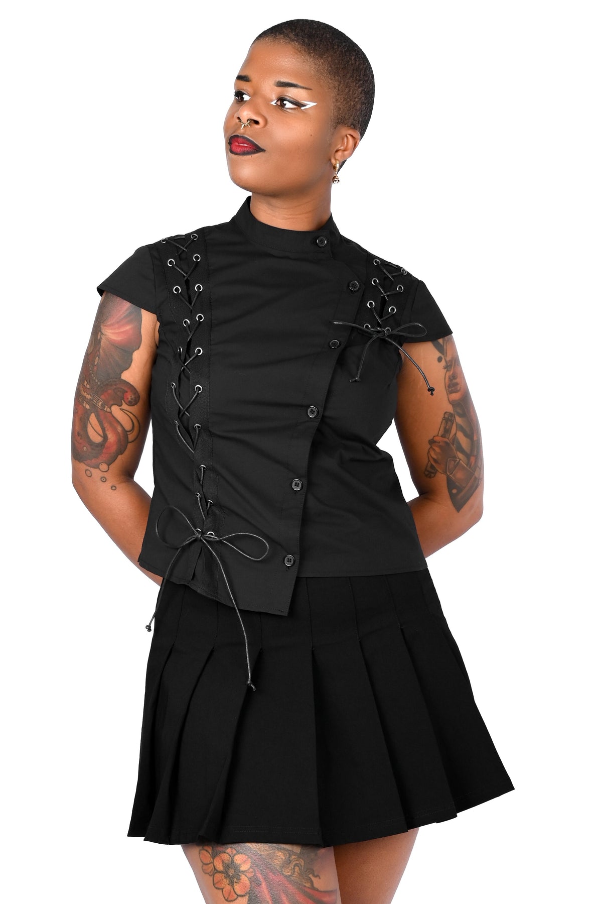 A model wearing a black short-sleeve mock-neck top with lace-up front details and a matching black pleated mini skirt, shown against a plain white background.