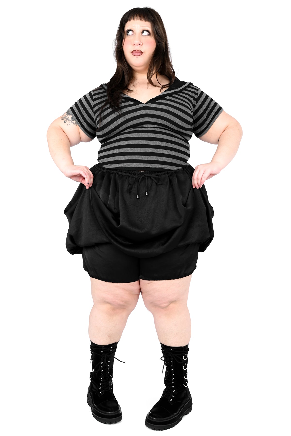 A plus-size model wearing a black and gray striped short-sleeve top, black balloon-style shorts, and black lace-up combat boots, standing against a white background.