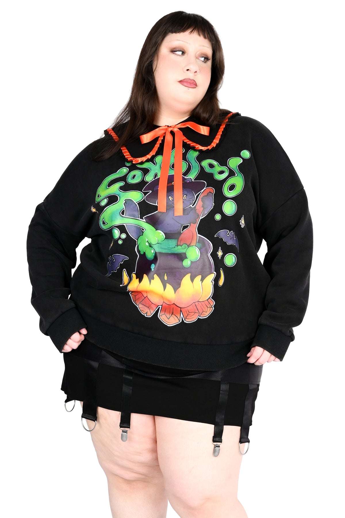A plus-size model wearing a black long-sleeve hoodie with a colorful graphic of a cauldron, green slime, and an orange collar with a bow, paired with a black mini skirt and garter straps.