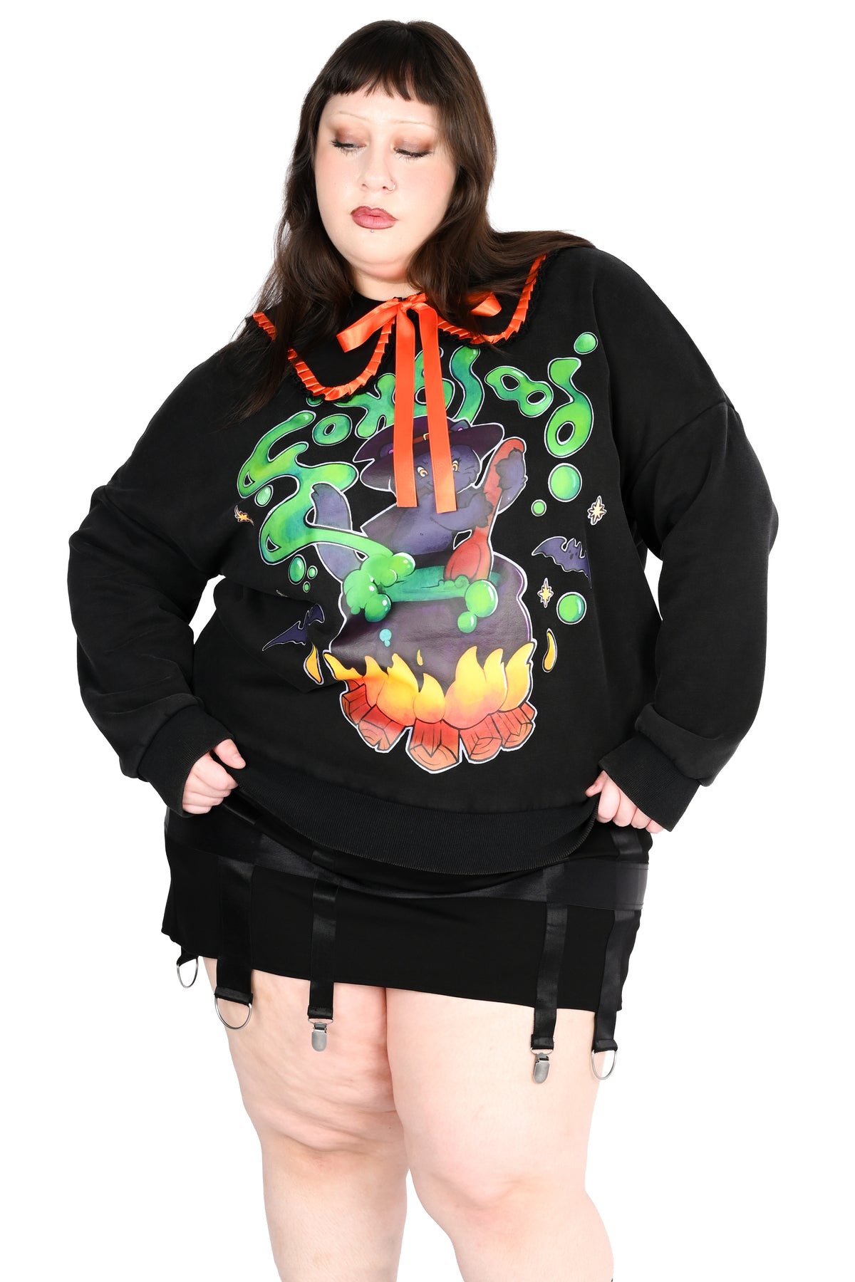 A plus-size model wearing a black sweater with a colorful 'Ghoulish' graphic on the front, featuring a cauldron, spider, and orange drawstring, paired with a black garter skirt.