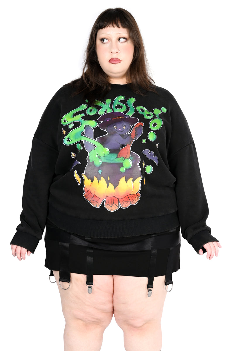 A model wearing a black crewneck sweatshirt with a colorful cat and cauldron graphic, styled with black garter shorts against a white background.