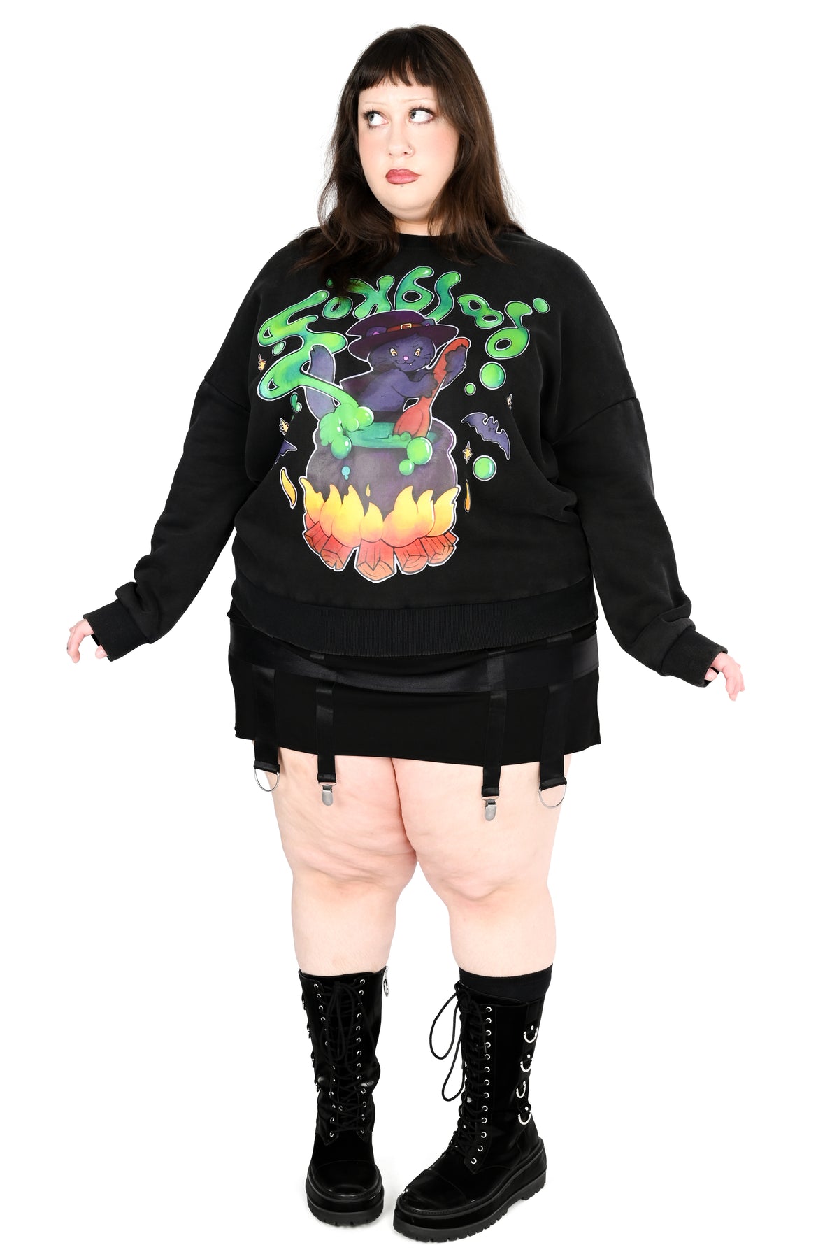 A plus-size model wearing a black long-sleeve sweatshirt with a colorful 'Monsters' graphic on the front, paired with a black mini skirt, garter belt, and black lace-up boots, against a solid white background.