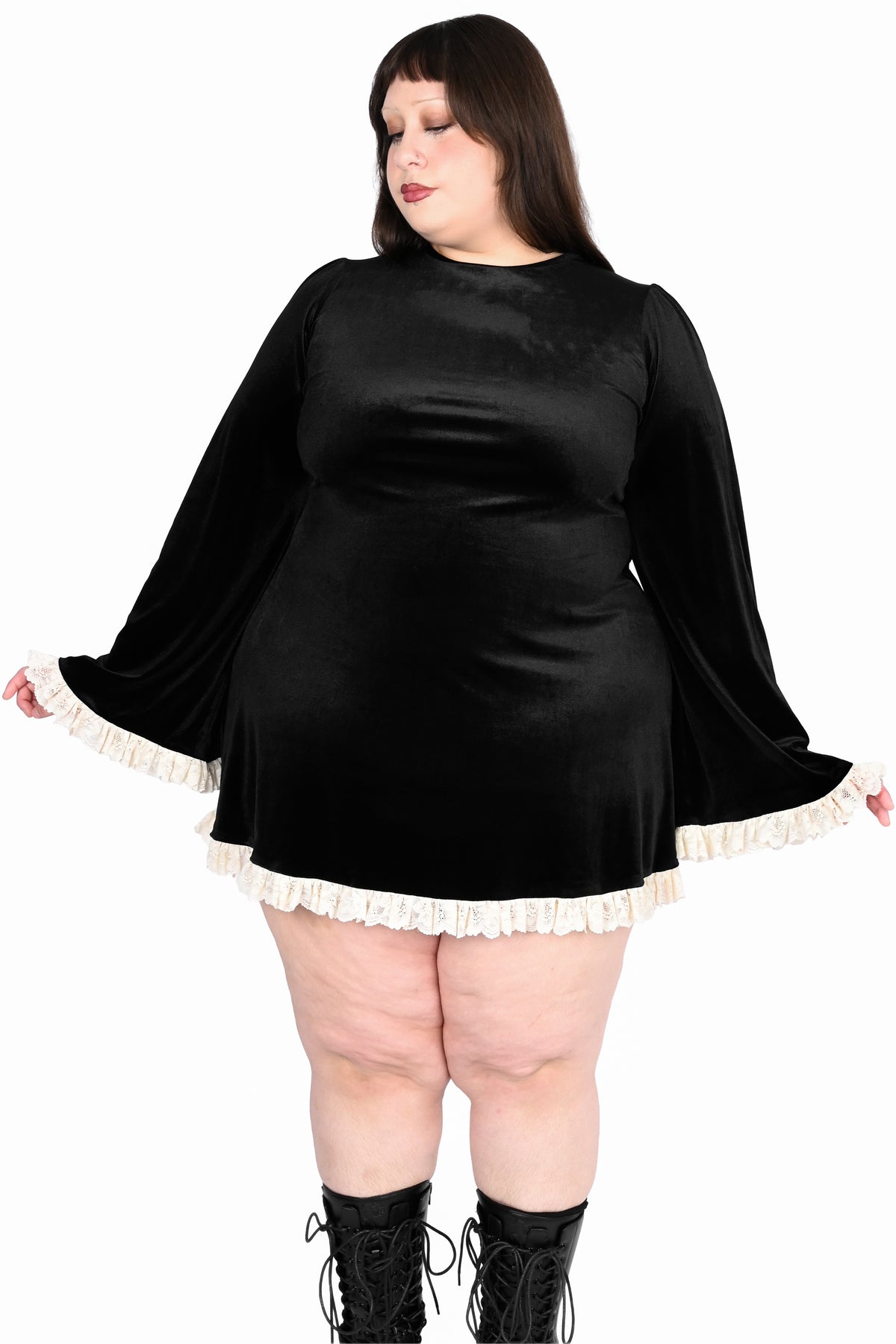 A plus-size model wearing a black velvet mini dress with dramatic bell sleeves and white ruffle trim, paired with black lace-up boots, against a plain white background.