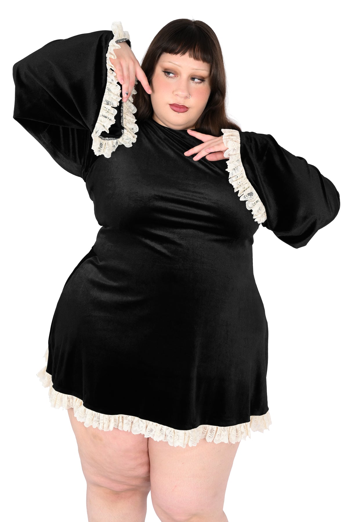 A plus-size woman modeling a black velvet mini dress with dramatic bell sleeves and cream lace trim, posed against a solid white background.