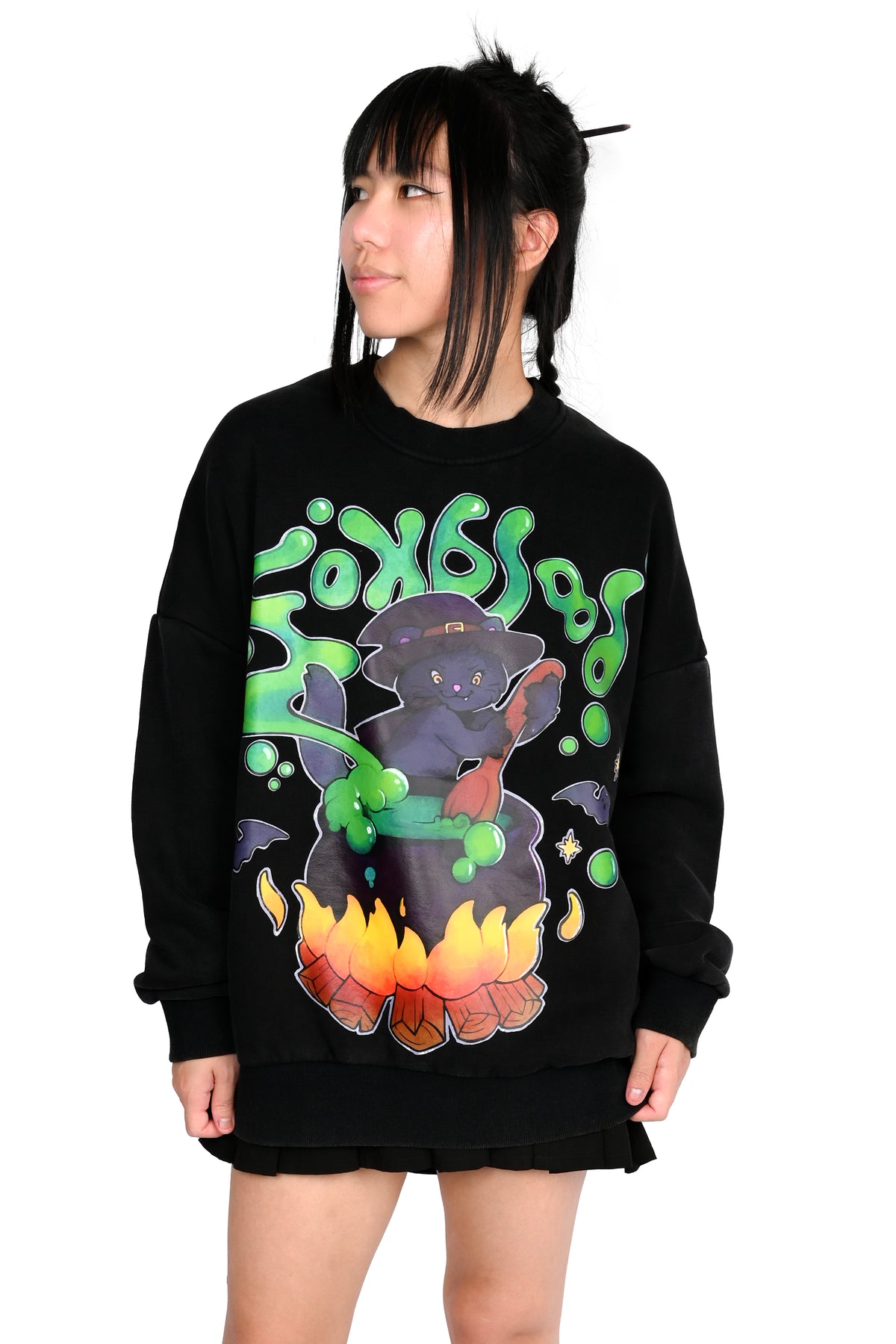 A model wearing a black crewneck sweatshirt featuring a colorful graphic of a wizard cat riding a broomstick with green smoke and the word 'FOXBLOOD' in green bubble letters.