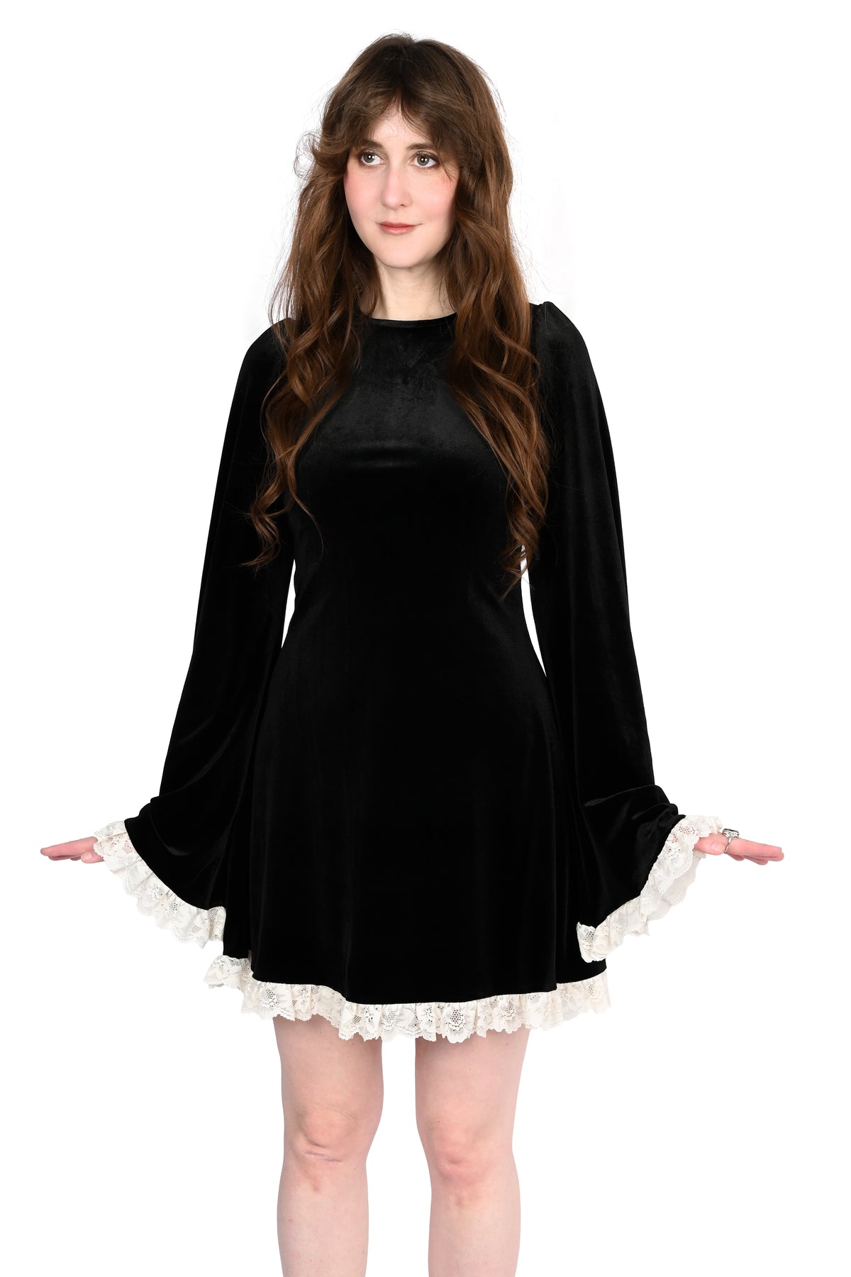 A woman modeling a black velvet mini dress with long bell sleeves and white lace trim at the cuffs and hem, set against a plain white background.
