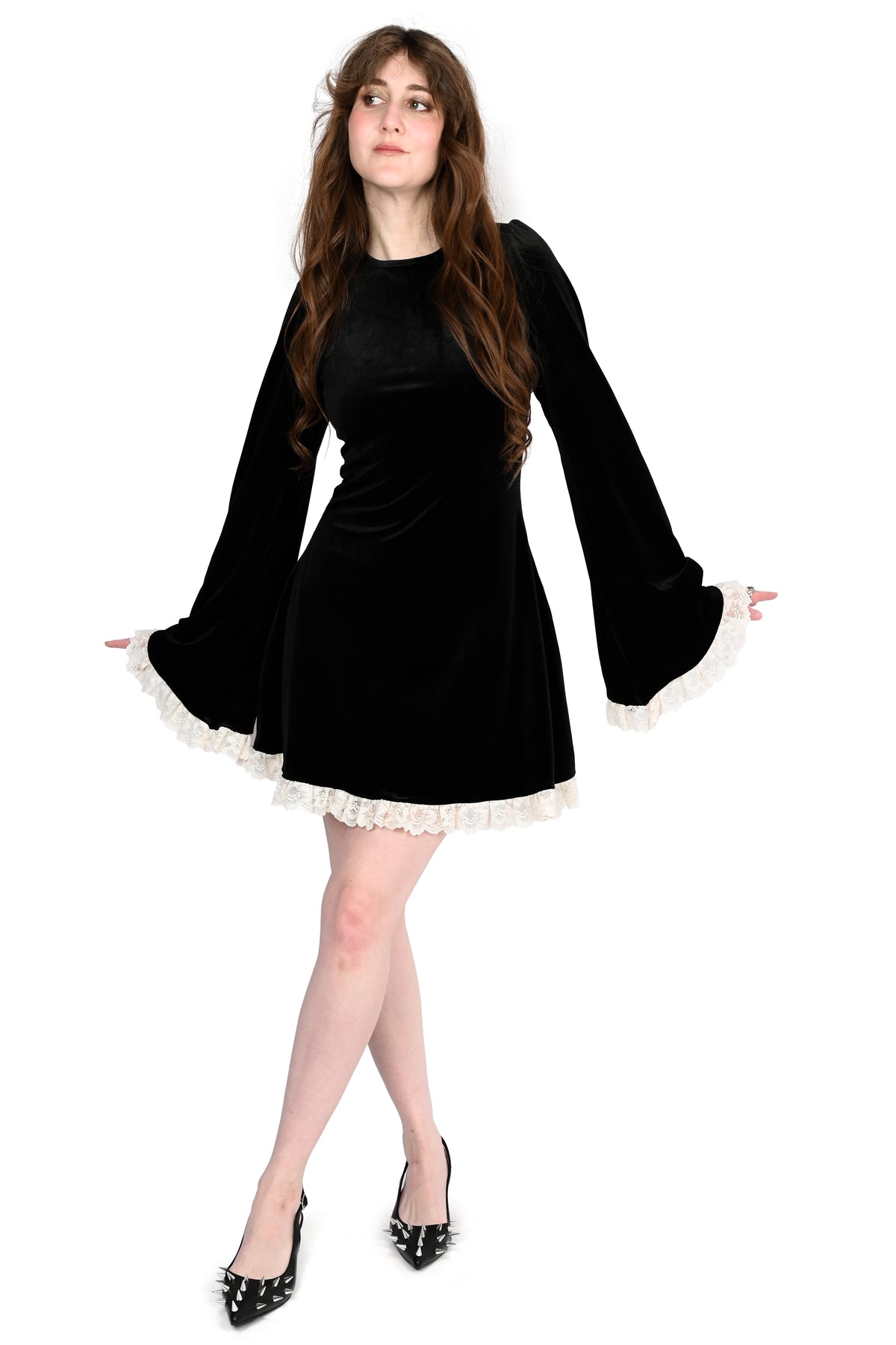A woman modeling a black velvet mini dress with long bell sleeves and white lace trim at the cuffs and hem, paired with black pointed-toe heels, against a plain white background.