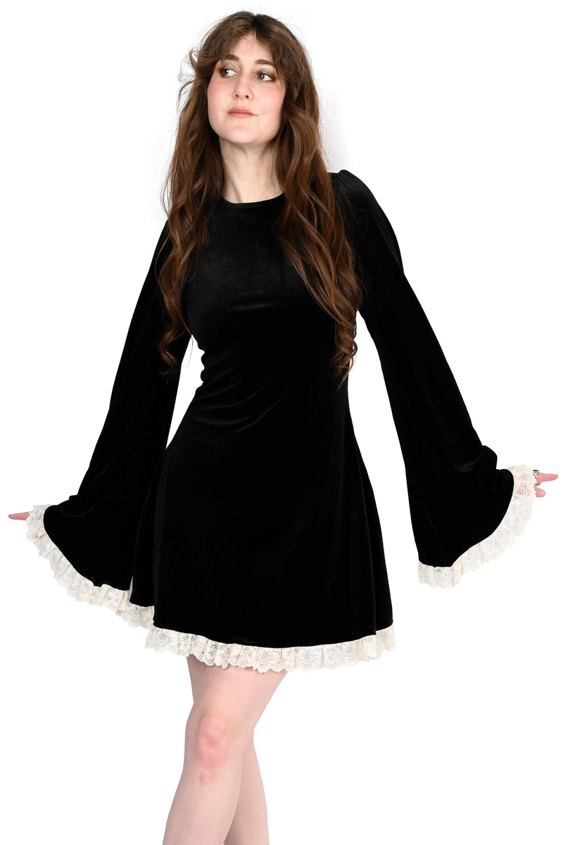 A woman modeling a black velvet mini dress with long bell sleeves and white lace trim at the cuffs and hem, paired with black pointed-toe heels, against a plain white background.