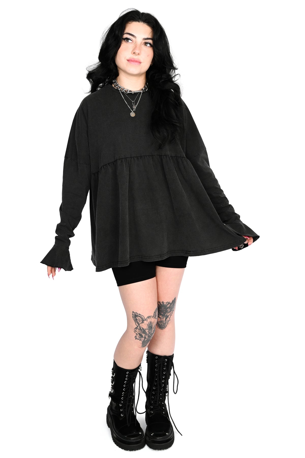 A model with long black hair is wearing a black long-sleeve tiered tunic top, black bike shorts, layered silver necklaces, and black lace-up combat boots, standing against a solid white background.