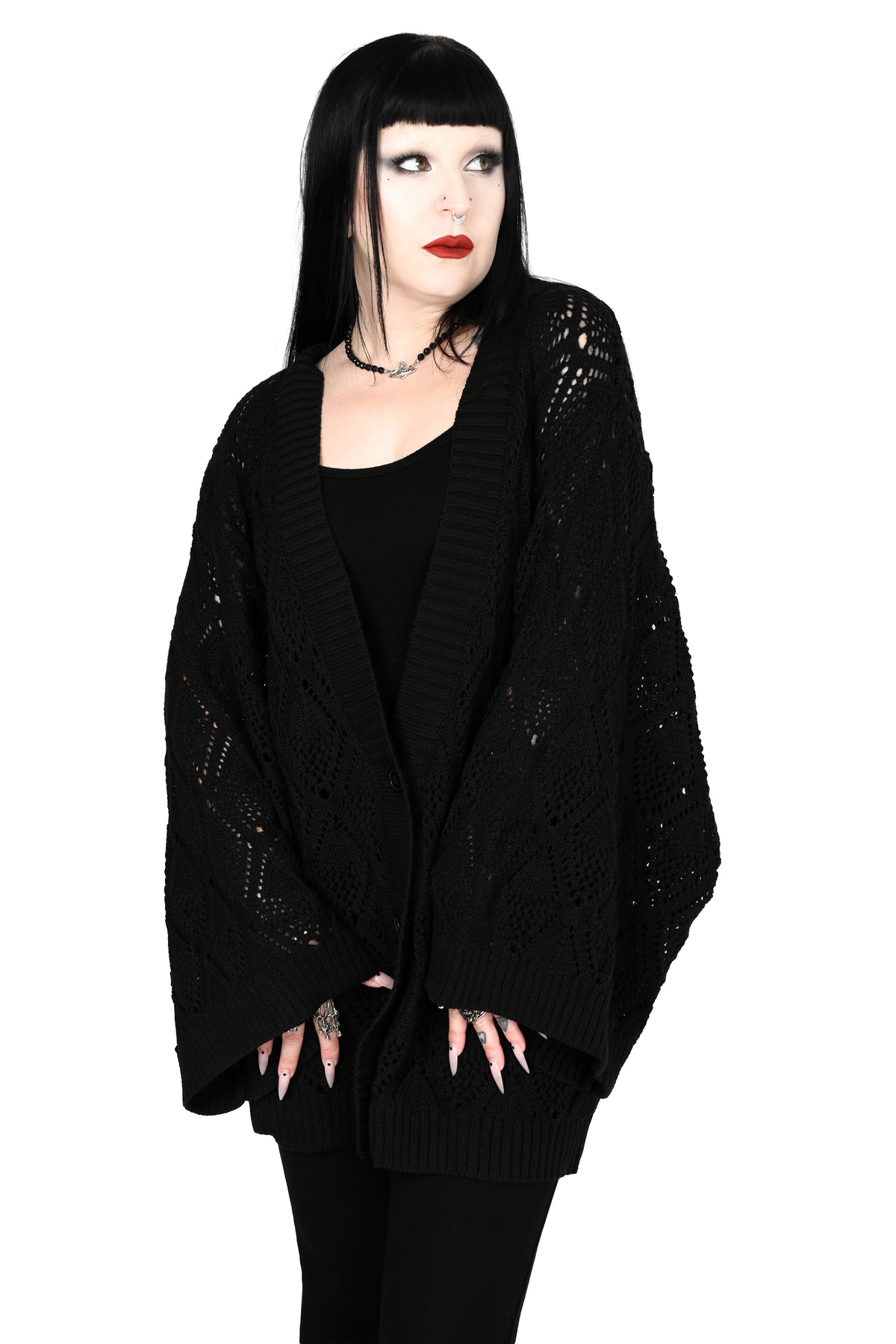 A model with black hair and bangs wearing an oversized black open-front knit cardigan with openwork lace sleeves, styled with a black top and black pants against a white background.