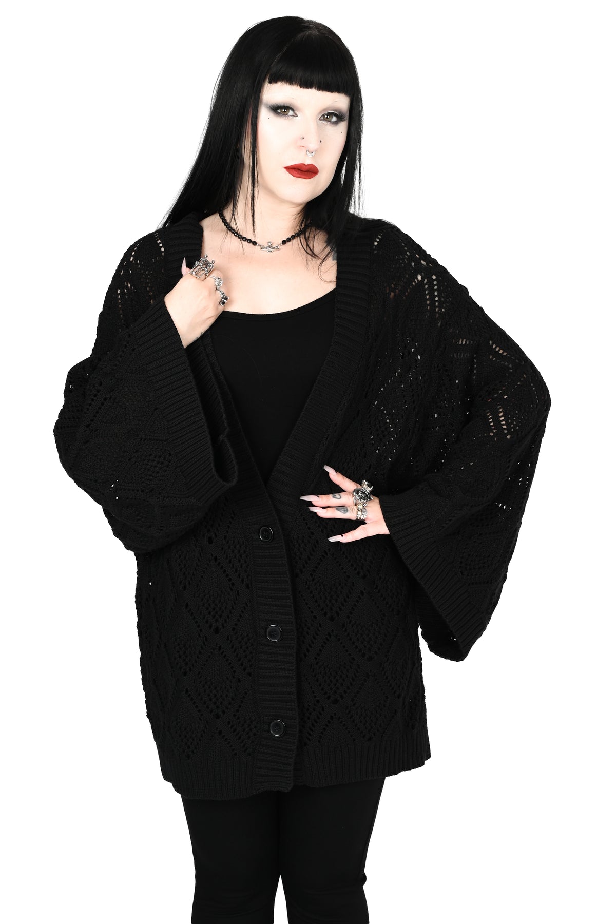 A model with long black hair and a gothic look wearing a black open-front cardigan with textured knit patterns, a black top, and black pants, set against a plain white background.