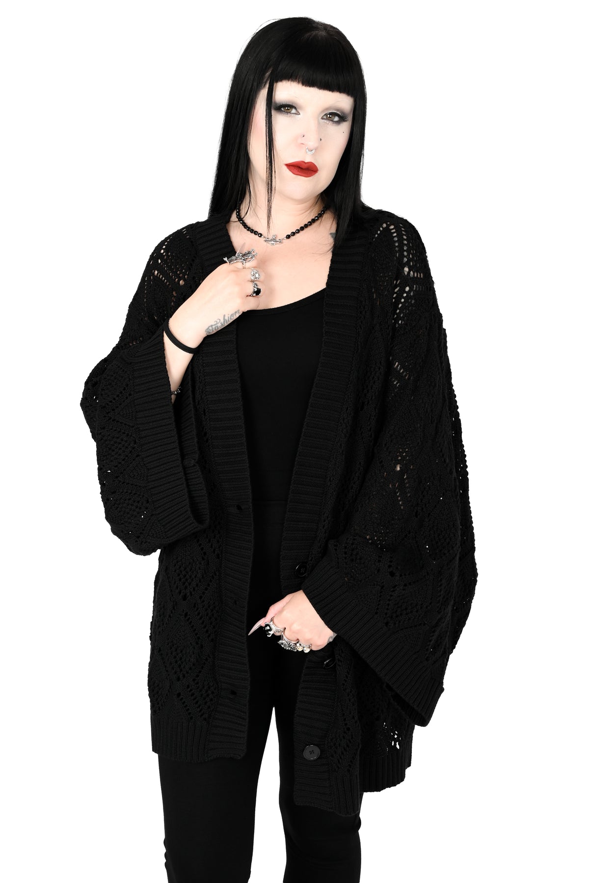 A model with black hair and bangs wearing a black open-front knit cardigan with textured openwork details, a black top, and black pants, posed against a solid white background.