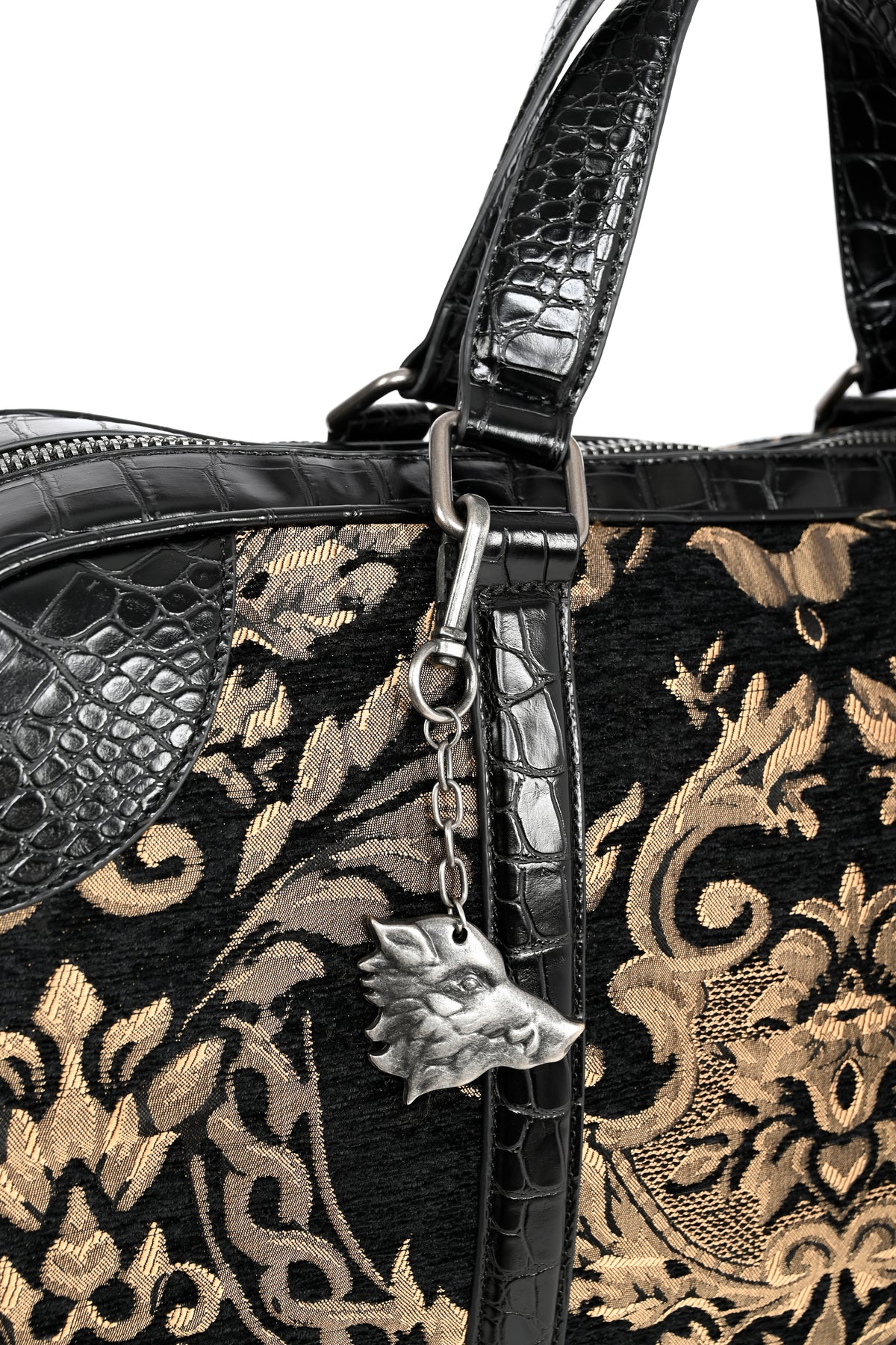Close-up of a black and gold damask-patterned handbag with textured black faux-croc trim, a silver-tone zipper pull, and a boar-head charm.