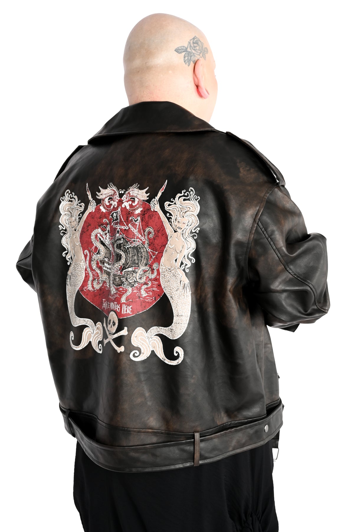 A model viewed from behind wearing a distressed black leather jacket with a large pirate themed graphic on the back, set against a plain white background. The graphic depicts two mermaids holding daggers on either side of a pirate ship that is being devoured by a kraken. There is a skull and crossbones under. The text 'there will be monsters' is visible