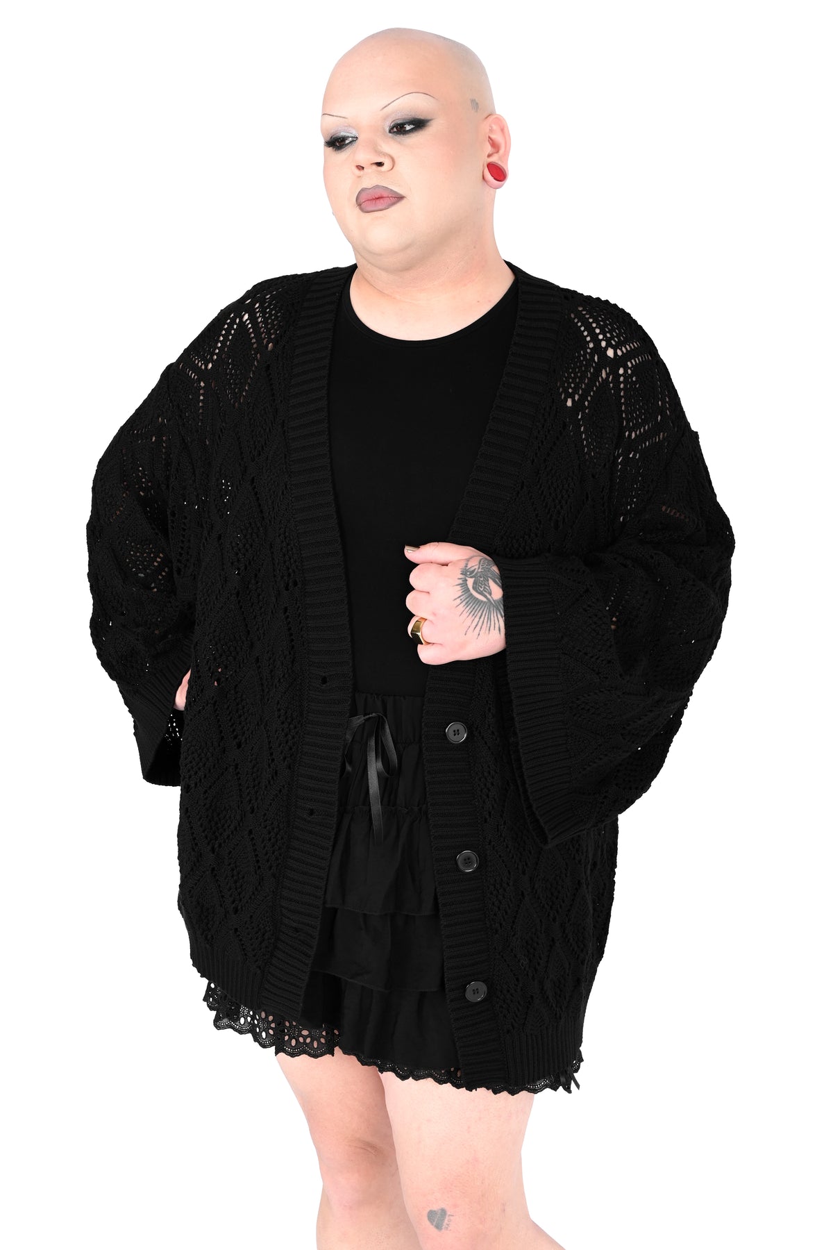 A plus-size model with a shaved head and dramatic makeup wearing a black open-front cardigan with textured knit details over a black outfit, set against a plain white background.