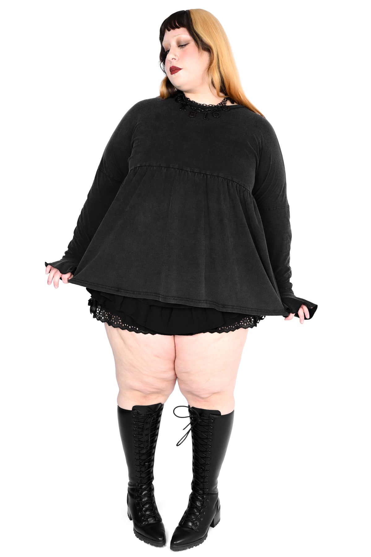 A plus-size model wearing a black long-sleeve top with a ruffled neckline, a matching black mini skirt with lace trim, and black lace-up knee-high boots, posed against a solid white background.
