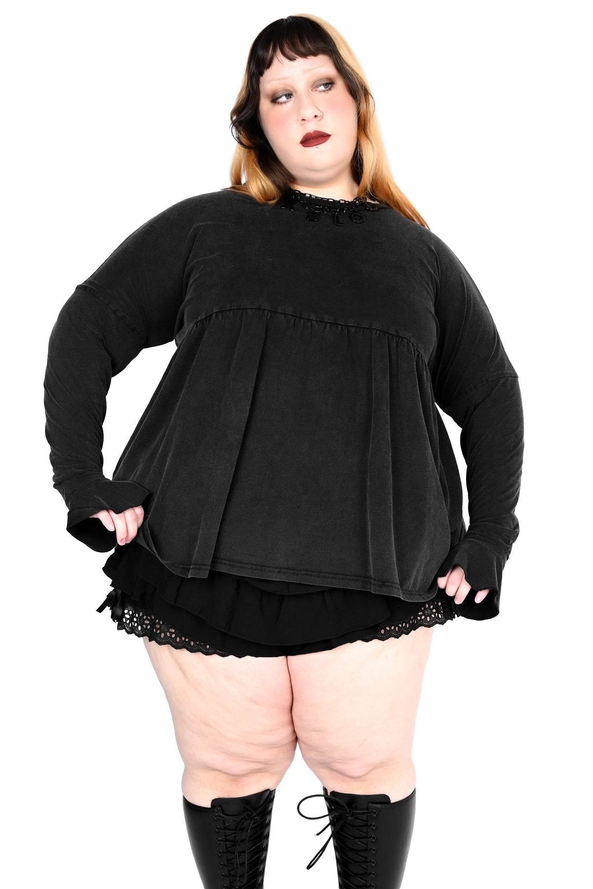 A plus-size model wearing a black long-sleeve top with a ruffled mock neck and layered peplum, paired with a black lace-trimmed mini skirt and black lace-up knee-high boots, against a plain white background.