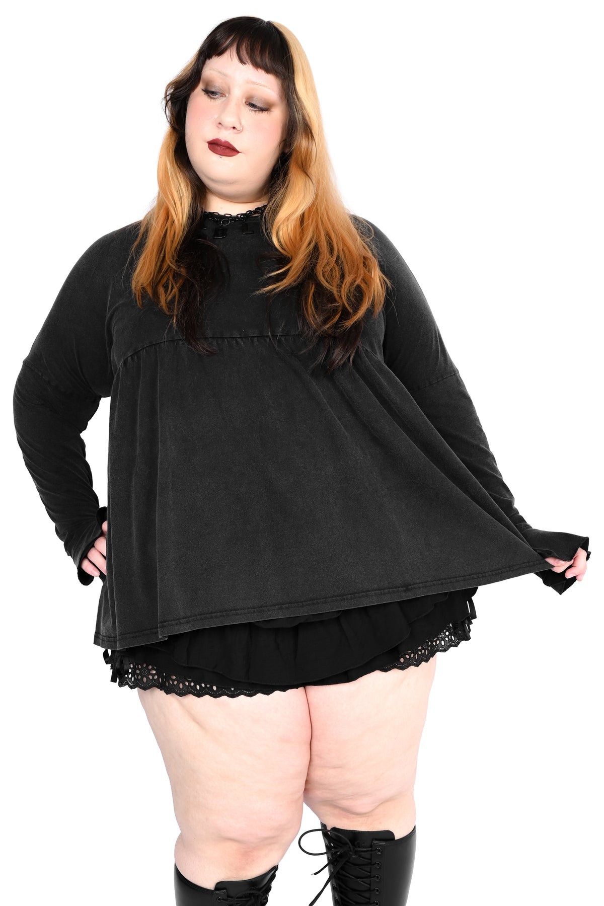 A plus-size model wearing a black long-sleeve mock-neck top with a ruffled hem, paired with a black ruffled skirt and black lace-up boots, against a solid white background.