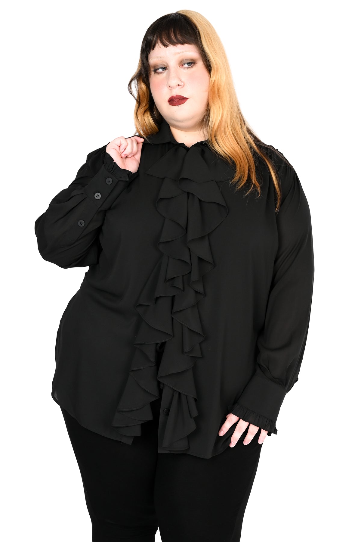 A plus-size model with blonde and dark hair wearing a black long-sleeve blouse with a dramatic ruffled front and black pants, posed against a solid white background.
