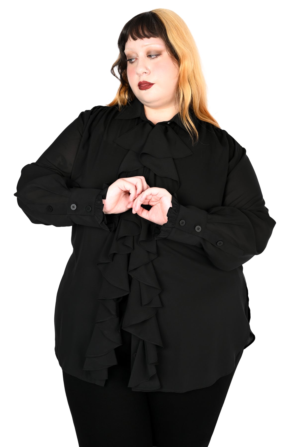 A woman modeling a black long-sleeve blouse with a high ruffled collar and cascading ruffle details down the front, paired with black pants, against a plain white background.