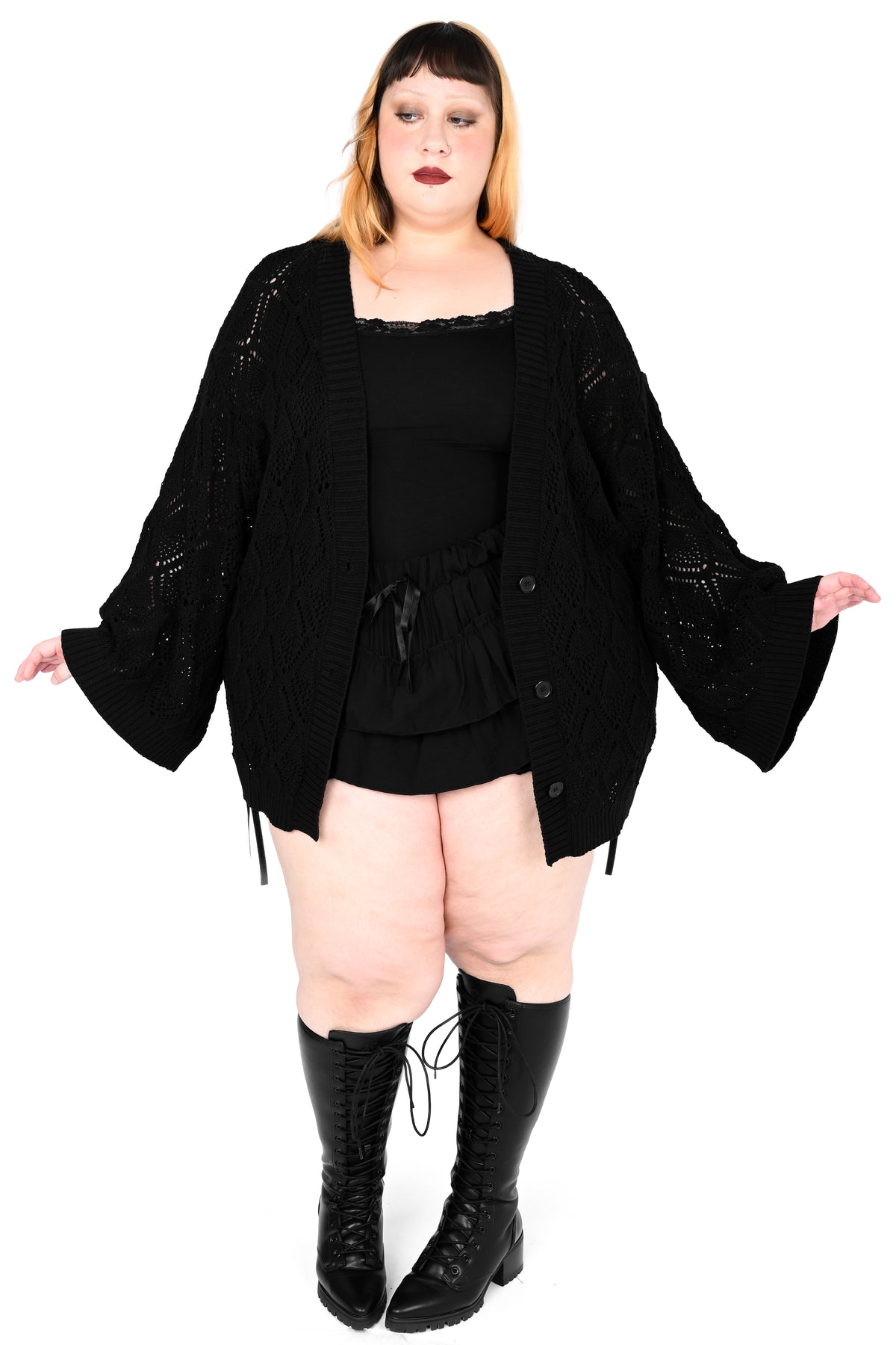 A plus-size model wearing a black knit cardigan with wide bell sleeves, a black top, a black skirt, and black lace-up boots, posed against a solid white background.