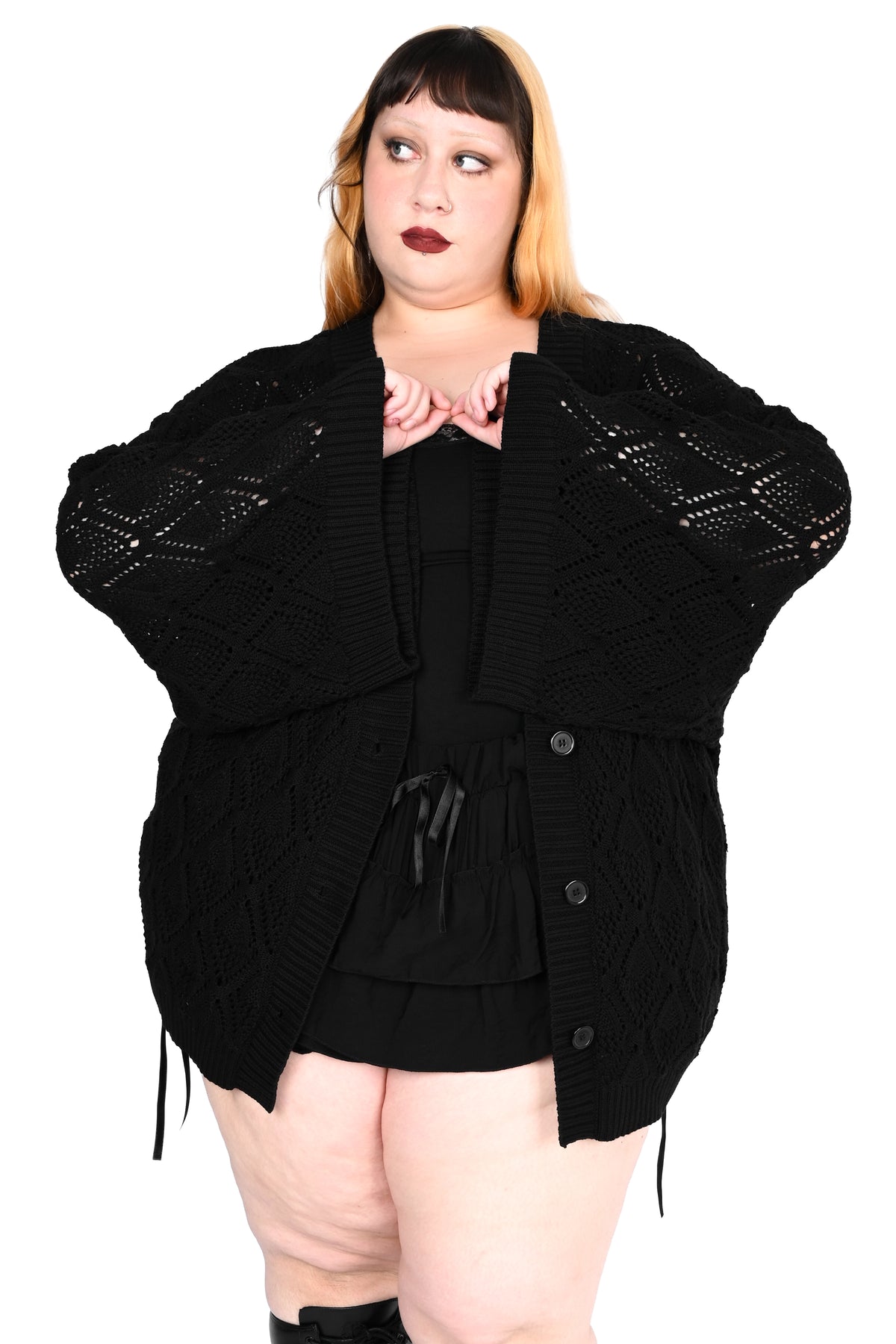A model wearing a black open-front knit cardigan with textured knit sleeves and drawstring details, styled over a black top and shorts, against a solid white background.