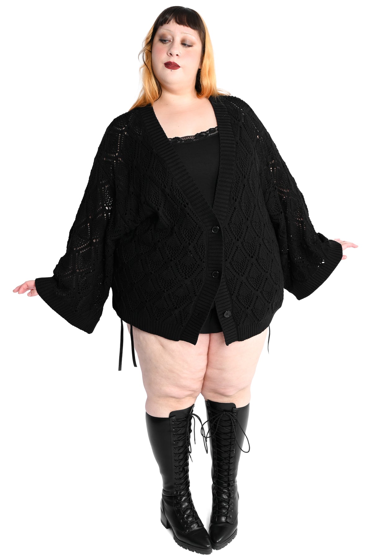 A plus-size model wearing a black open-front knit cardigan with wide bell sleeves, a black top, and black lace-up knee-high boots, posed against a solid white background.