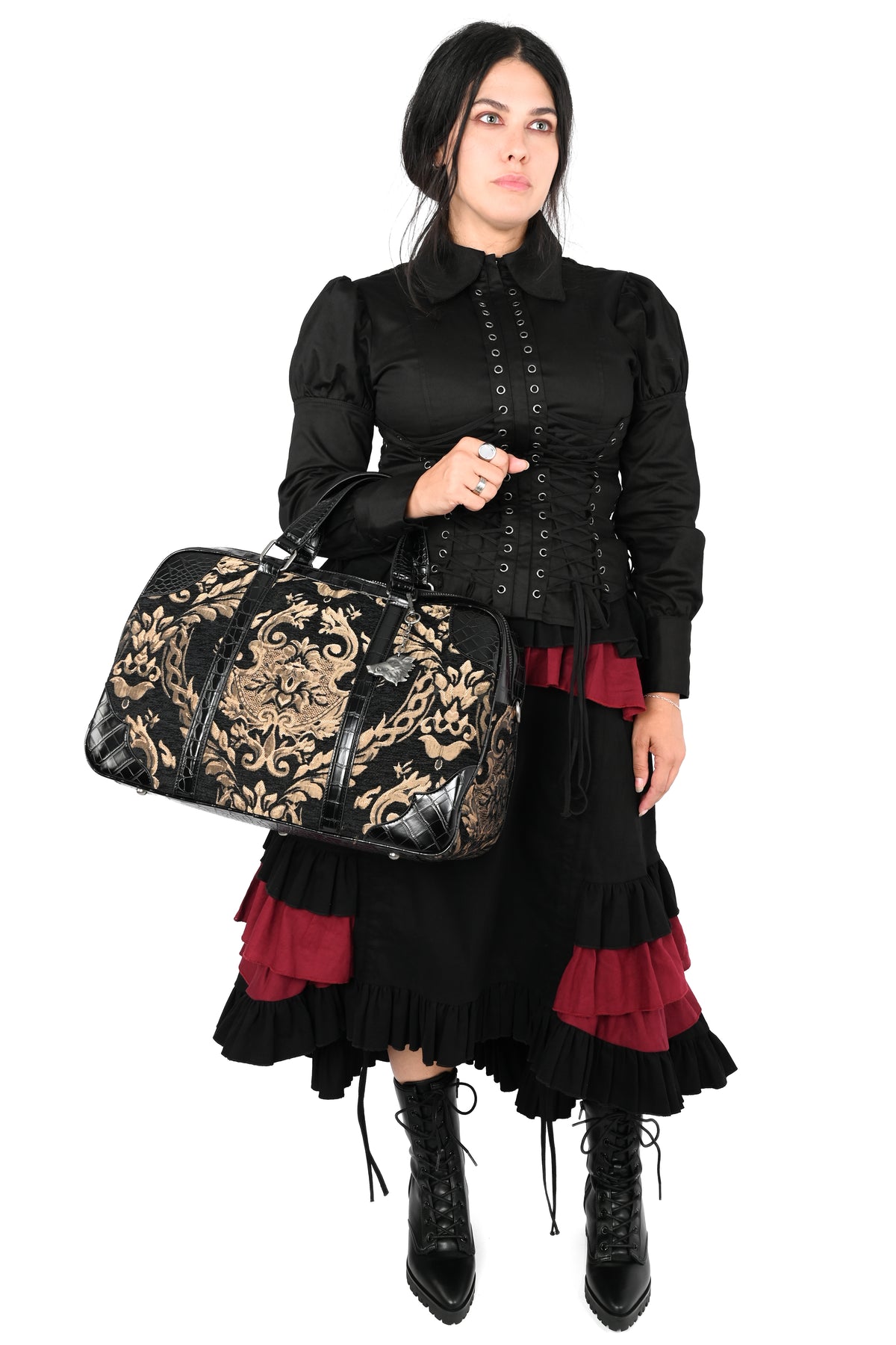 A woman modeling a black gothic-style outfit with a ruffled skirt, holding a black and gold patterned duffel bag against a plain white background.