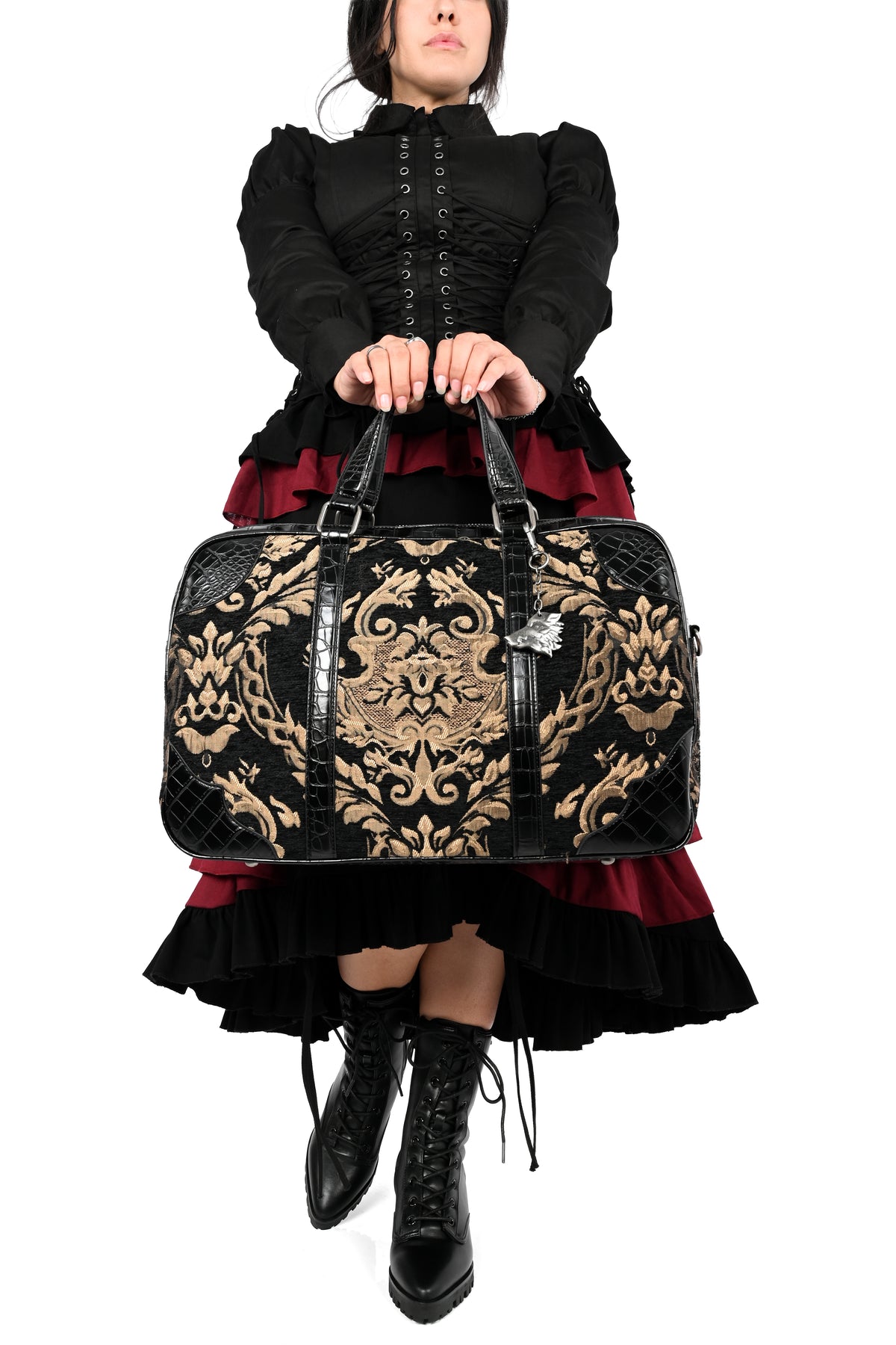 A model wearing a black and dark red gothic outfit holds a large black duffel bag with an ornate gold pattern and black faux-leather handles, set against a plain white background.