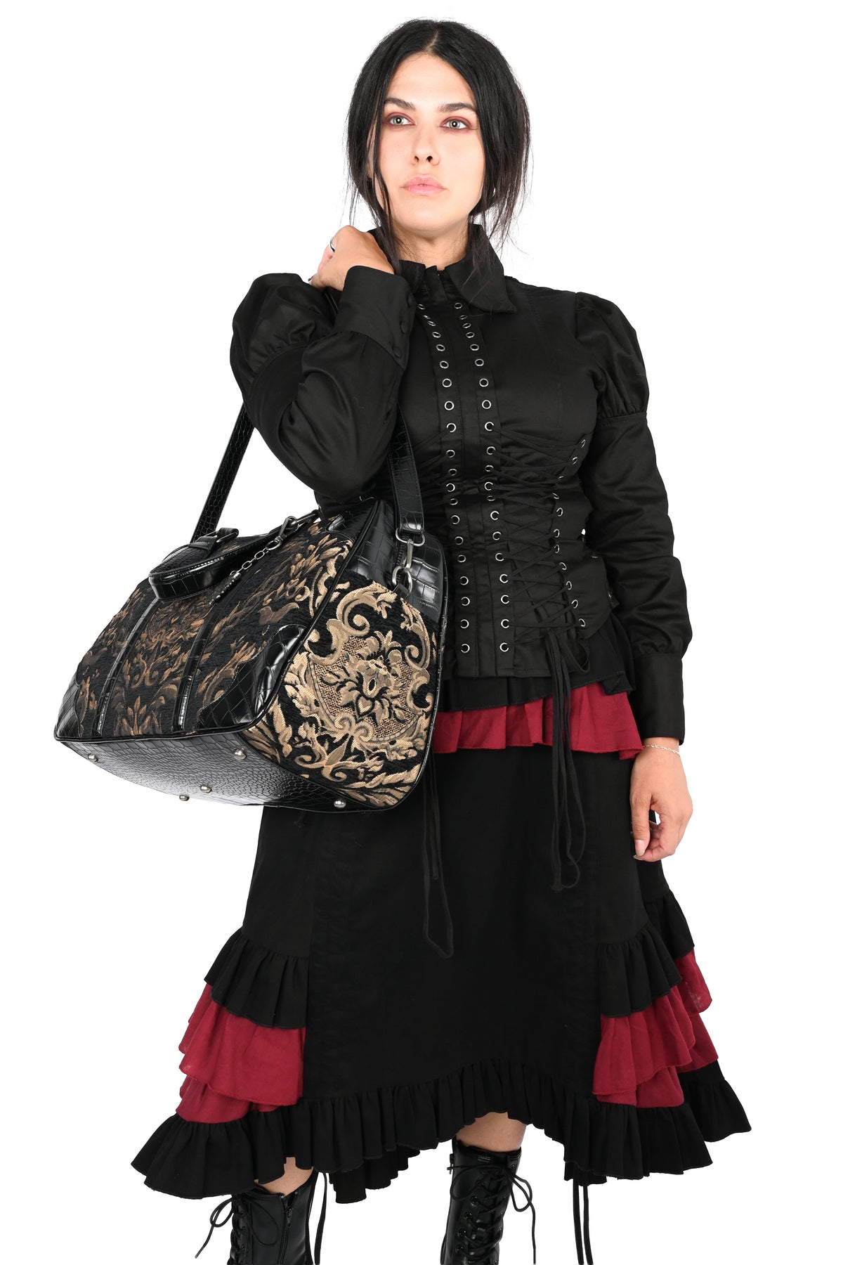 A woman modeling a black gothic-style outfit with red ruffle accents, carrying a black and gold damask-patterned shoulder bag against a plain white background.