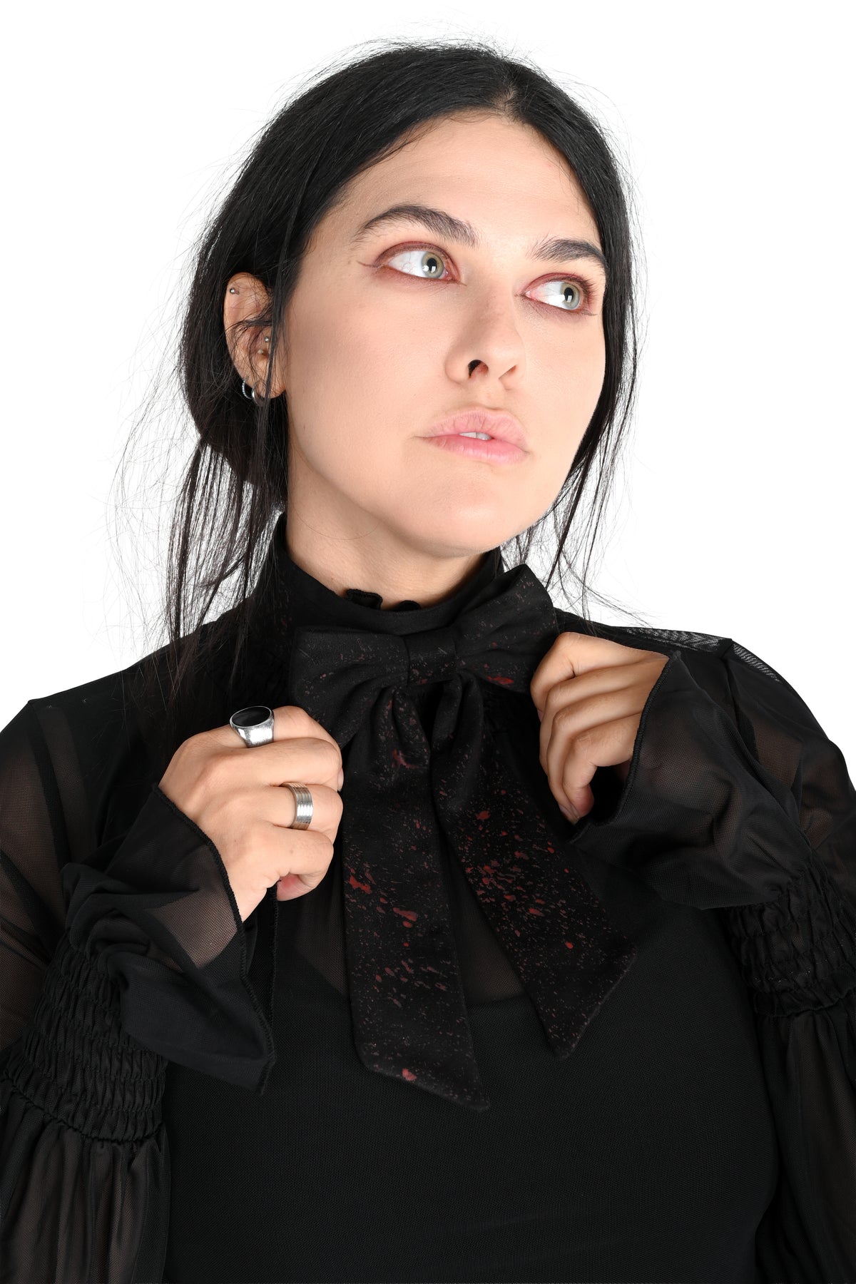 A woman with dark hair looking upward and to the side, wearing a black sheer long-sleeve blouse showing off a wide black choker with giant bow that has a red splatter over it. set against a plain white background.