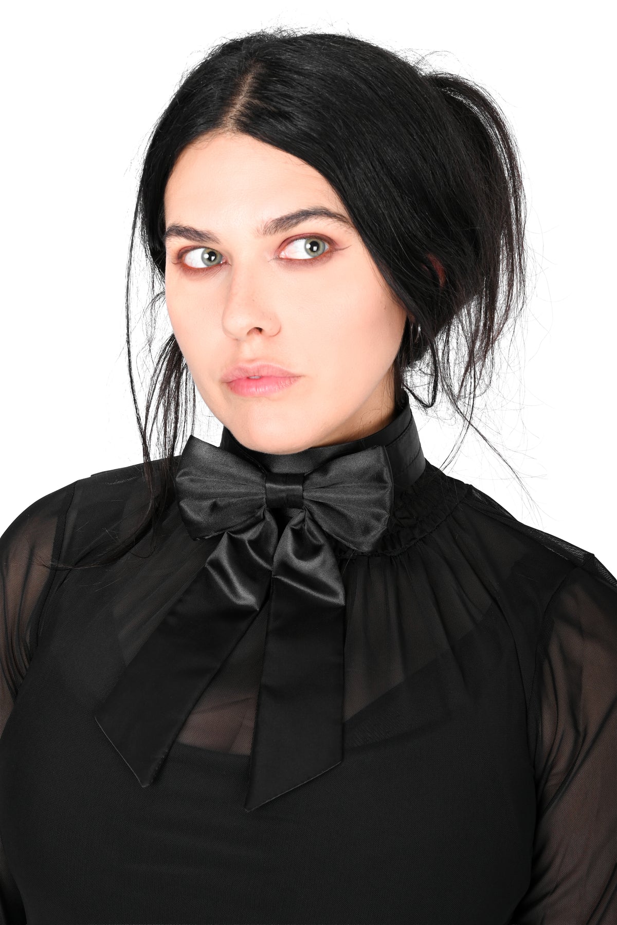 A woman with dark hair wearing a black sheer long-sleeve blouse showing off a choker satin bow with long tails photographed against a plain white background.