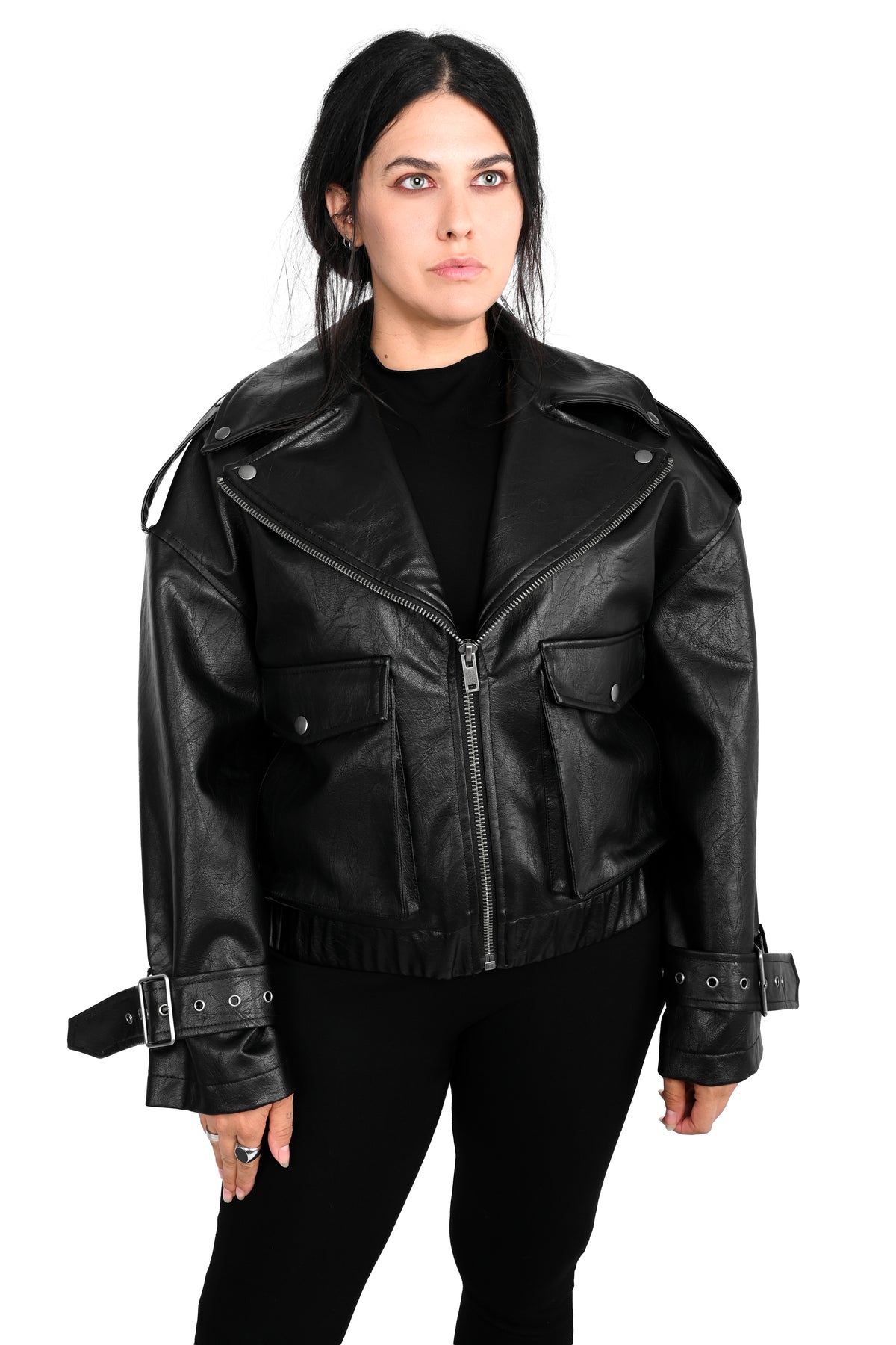 A woman modeling a black faux leather moto jacket with a front zipper, flap chest pockets, and adjustable cuff straps, paired with a black turtleneck and black pants against a white background.