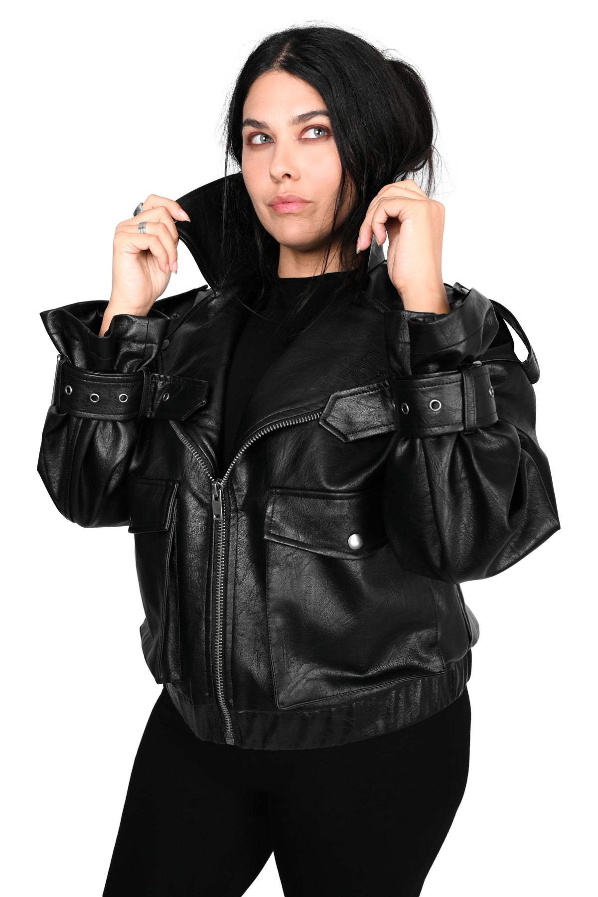 A woman modeling a black faux-leather moto jacket with unique gathered sleeve details and a front zipper, paired with black pants against a solid white background.