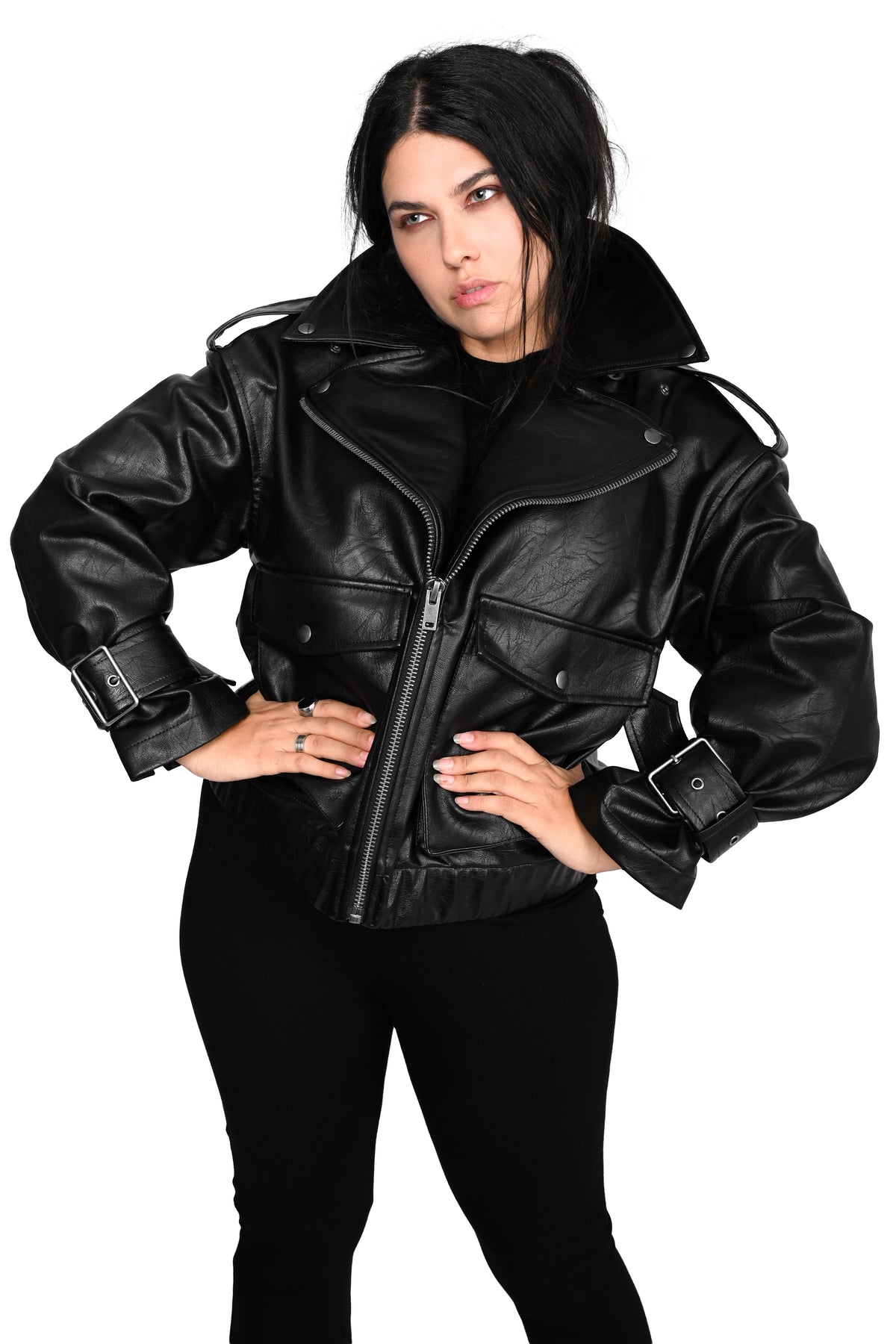 A woman modeling a black faux-leather biker jacket with a high collar, front zipper, and adjustable cuff straps, paired with black pants against a plain white background.