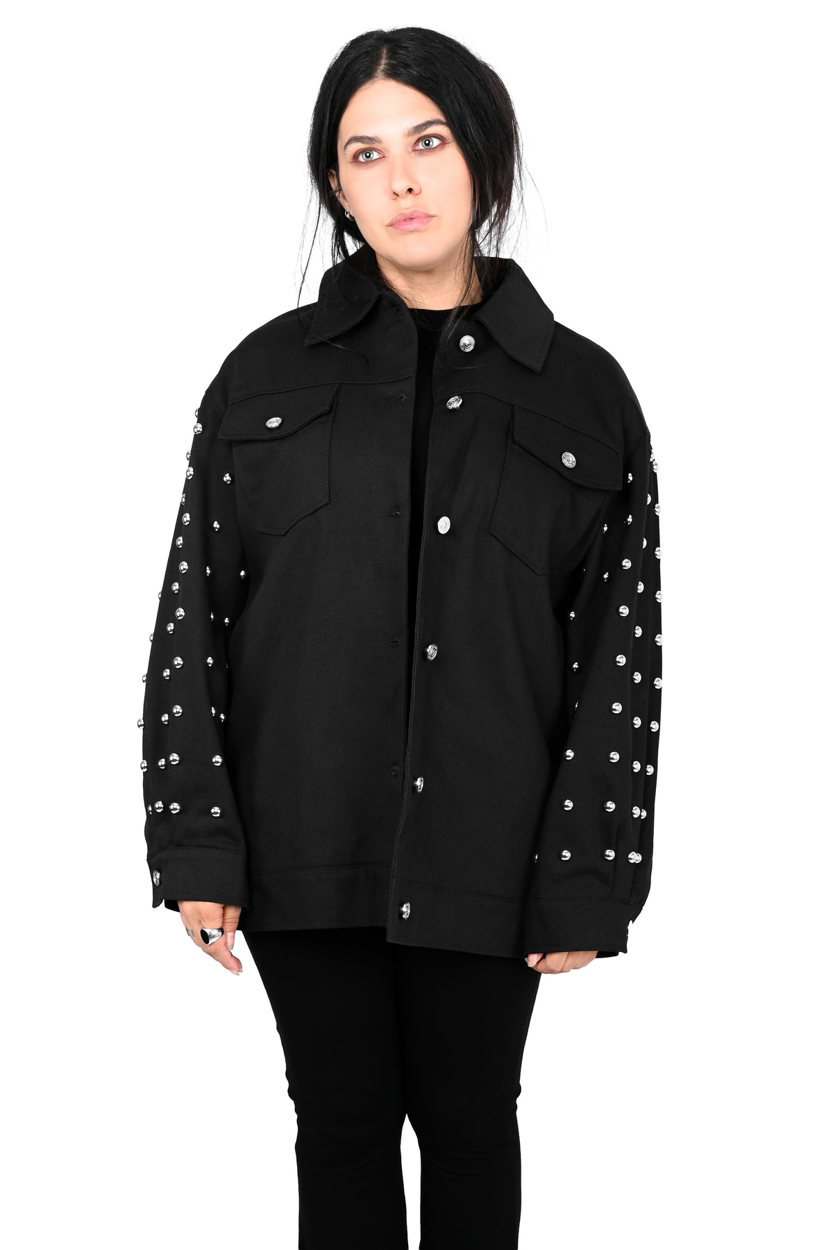 A model wearing a black denim jacket with silver studs on the sleeves, paired with black pants, photographed against a solid white background.