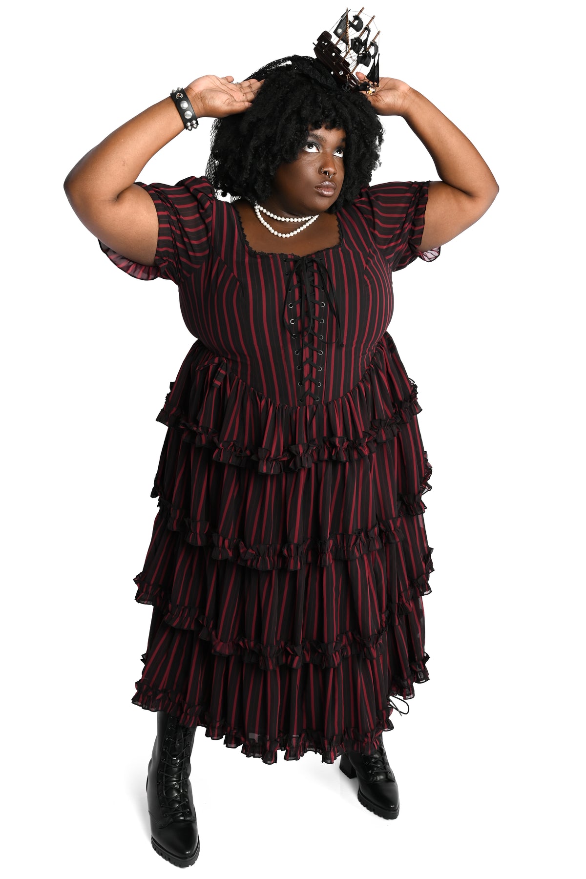 A plus sized model wearing a black and red vertically striped tiered ruffle dress with a lace-up front, a fascinator with a ship on it, and black boots, set against a plain white background.