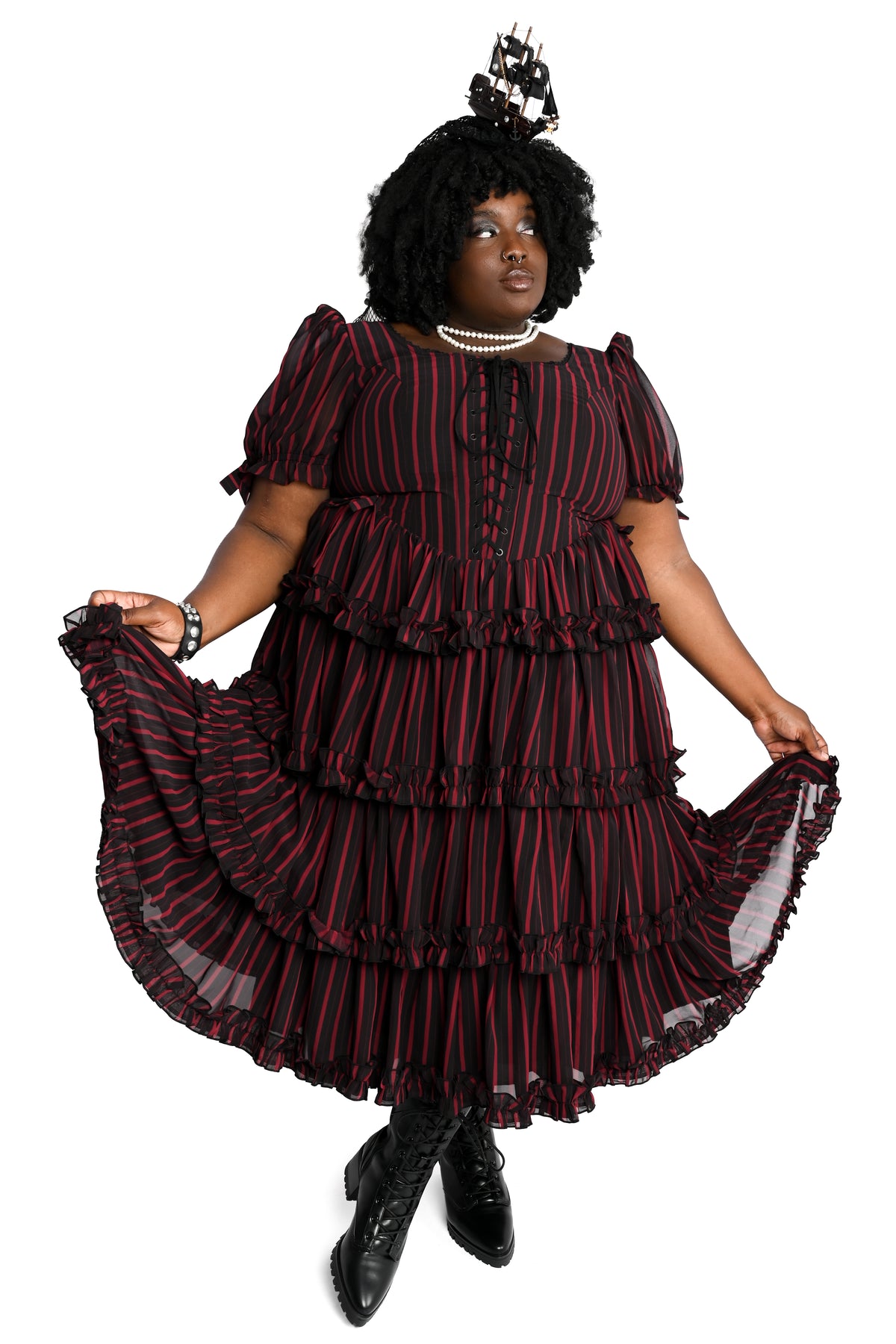 A woman modeling a black and red vertically striped, tiered ruffle dress with puff sleeves, holding out the sides of the skirt to show its volume, against a plain white background.