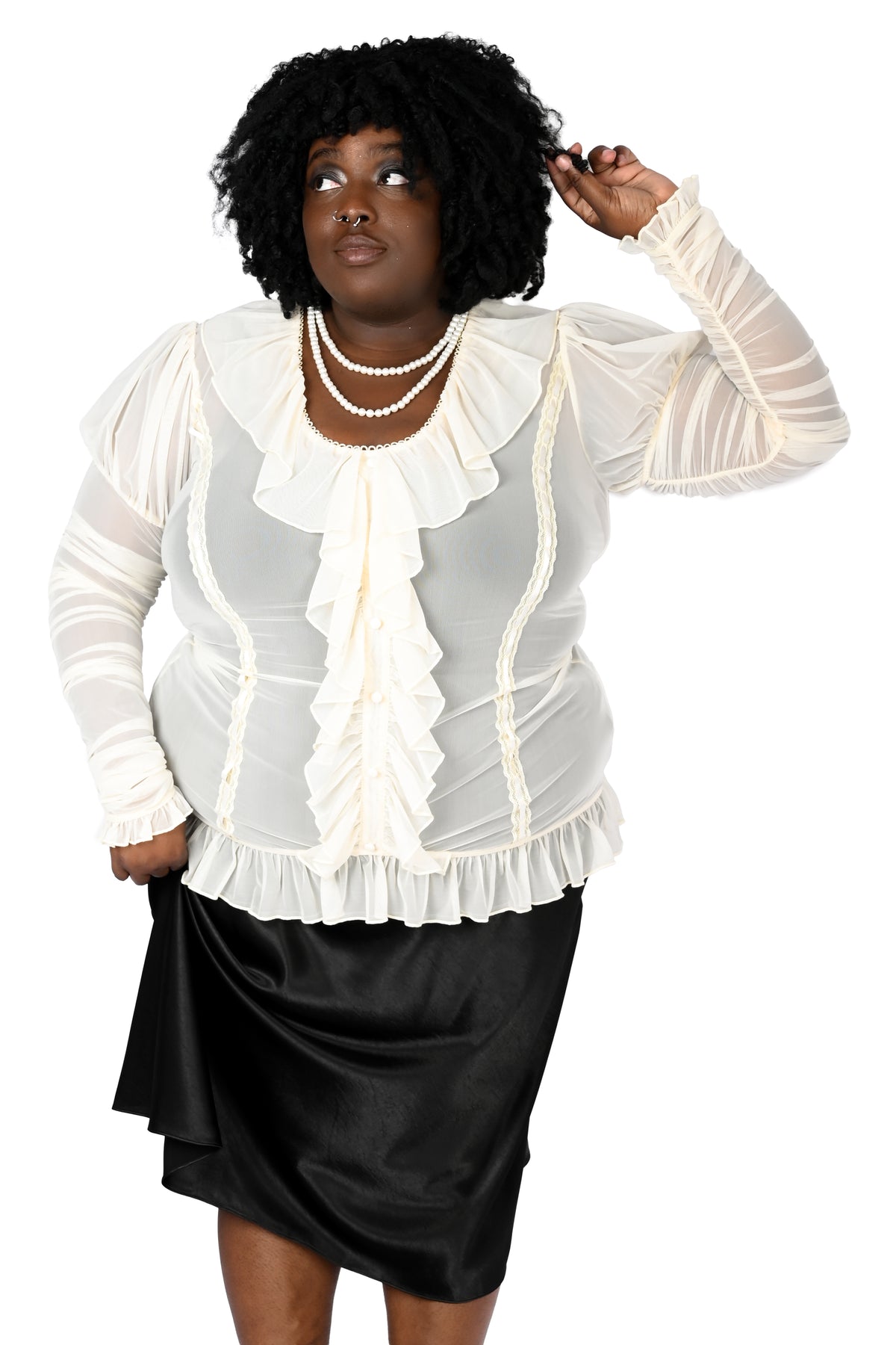 A woman modeling a cream ruffled long-sleeve blouse with sheer sleeves and a black satin skirt against a plain white background.
