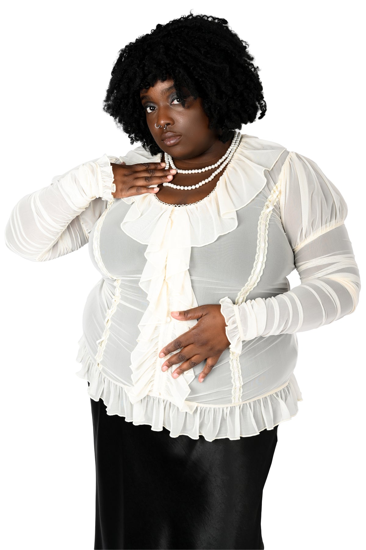 A woman modeling a white ruffled long-sleeve blouse with sheer mesh sleeves and a pleated peplum-style hem, paired with black pants and pearl necklaces, against a plain white background.