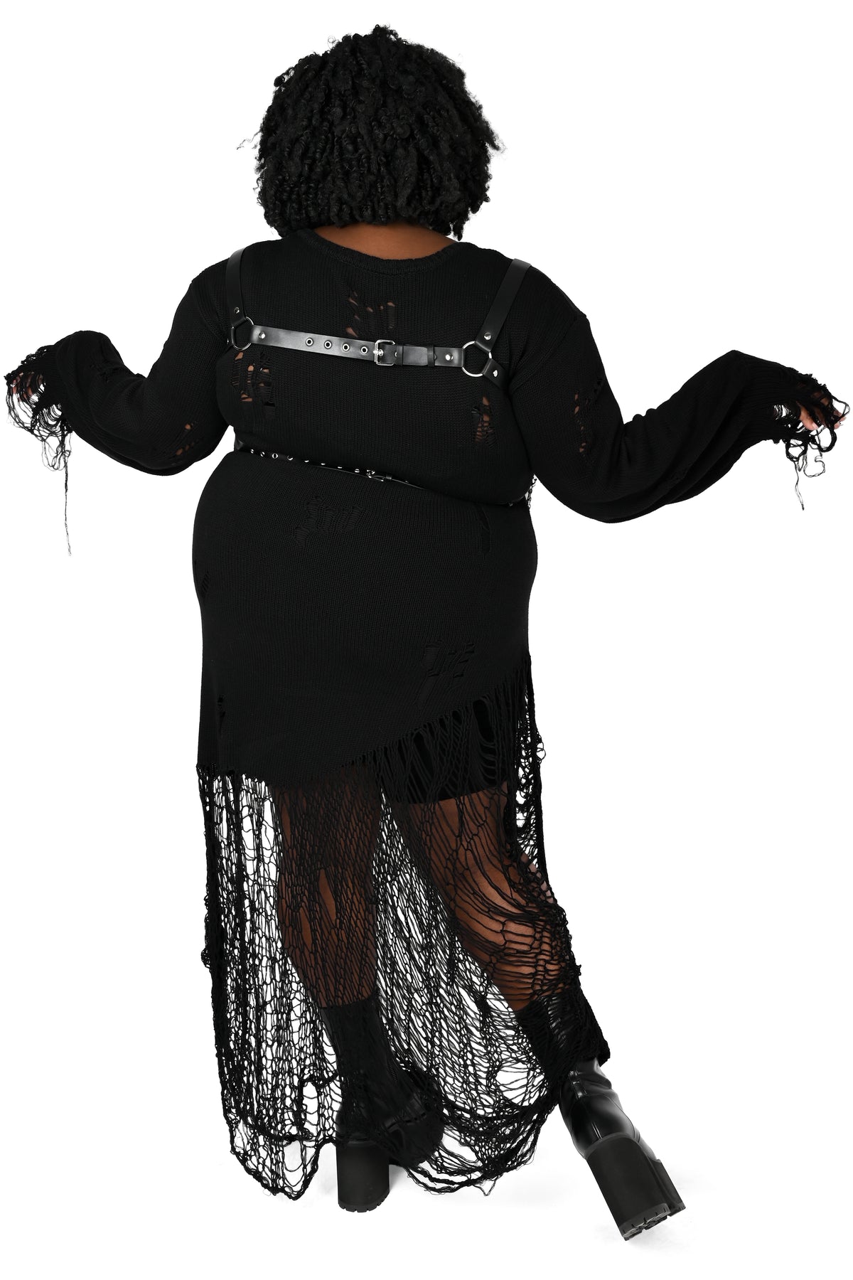 A person shown from the back wearing a black distressed long-sleeve dress with fringe, a metallic harness-style strap across the upper back, and sheer black tights, against a plain white background.