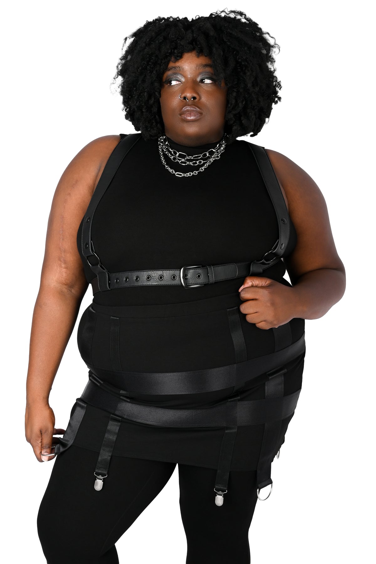 A plus-size model wearing a black harness over a sleeveless top paired with a skirt, posed against a plain white background.
