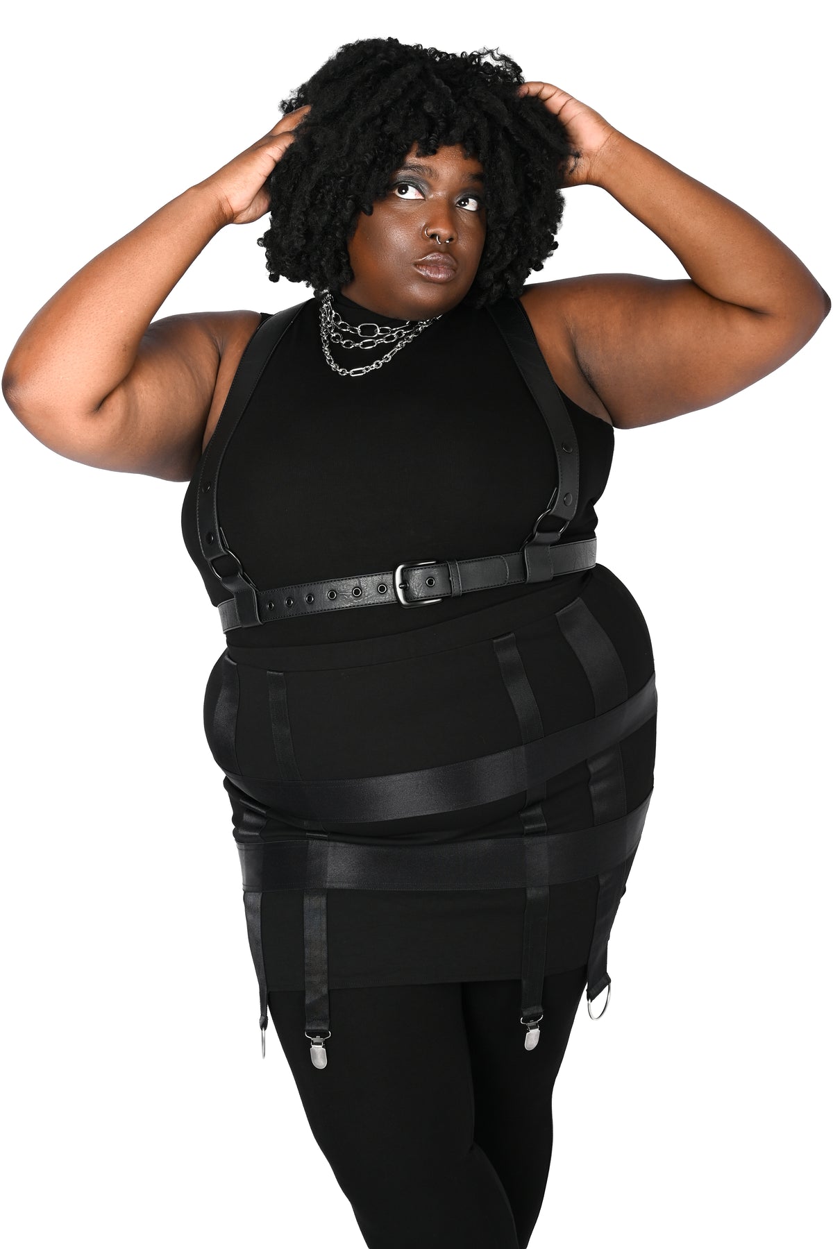 A plus-size model wearing a black harness with a skirt and garter details, styled with black leggings and layered silver necklaces, set against a plain white background.