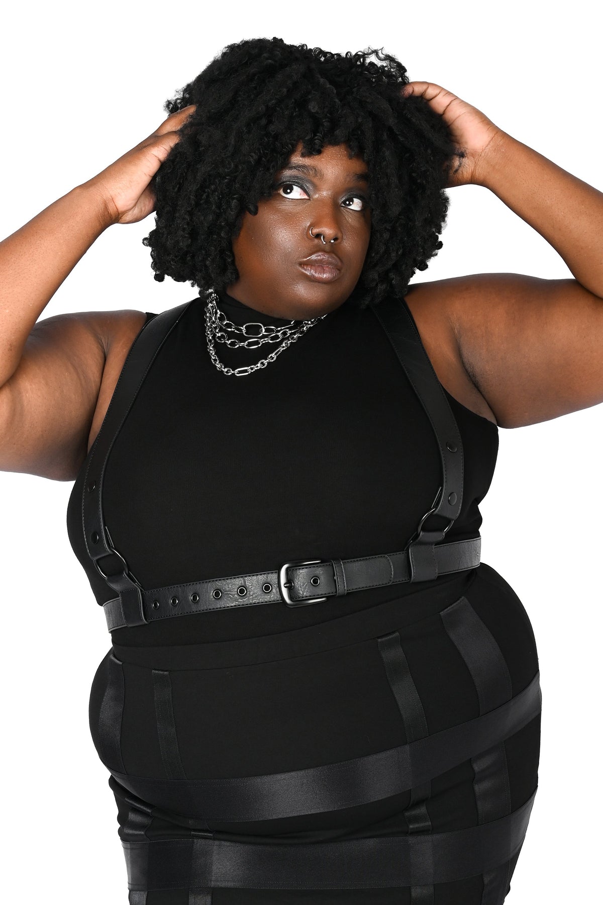 A plus-size model wearing a black harness, styled with black leggings and layered silver necklaces, set against a plain white background.