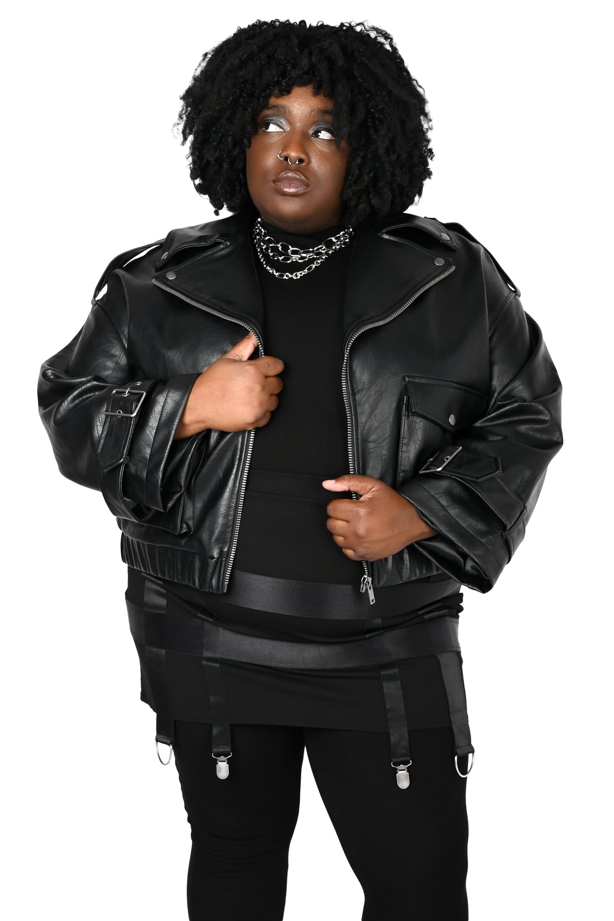 A person modeling a black faux-leather moto jacket with silver zippers and snaps, worn over a black top and black pants with suspenders, on a plain white background.
