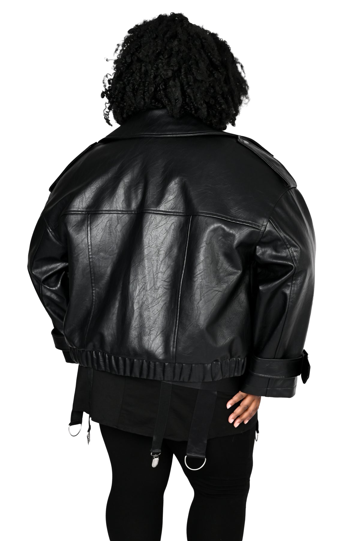 A person shown from the back wearing an oversized black leather jacket with shoulder epaulets and a ruffled hem, paired with black pants, against a plain white background.