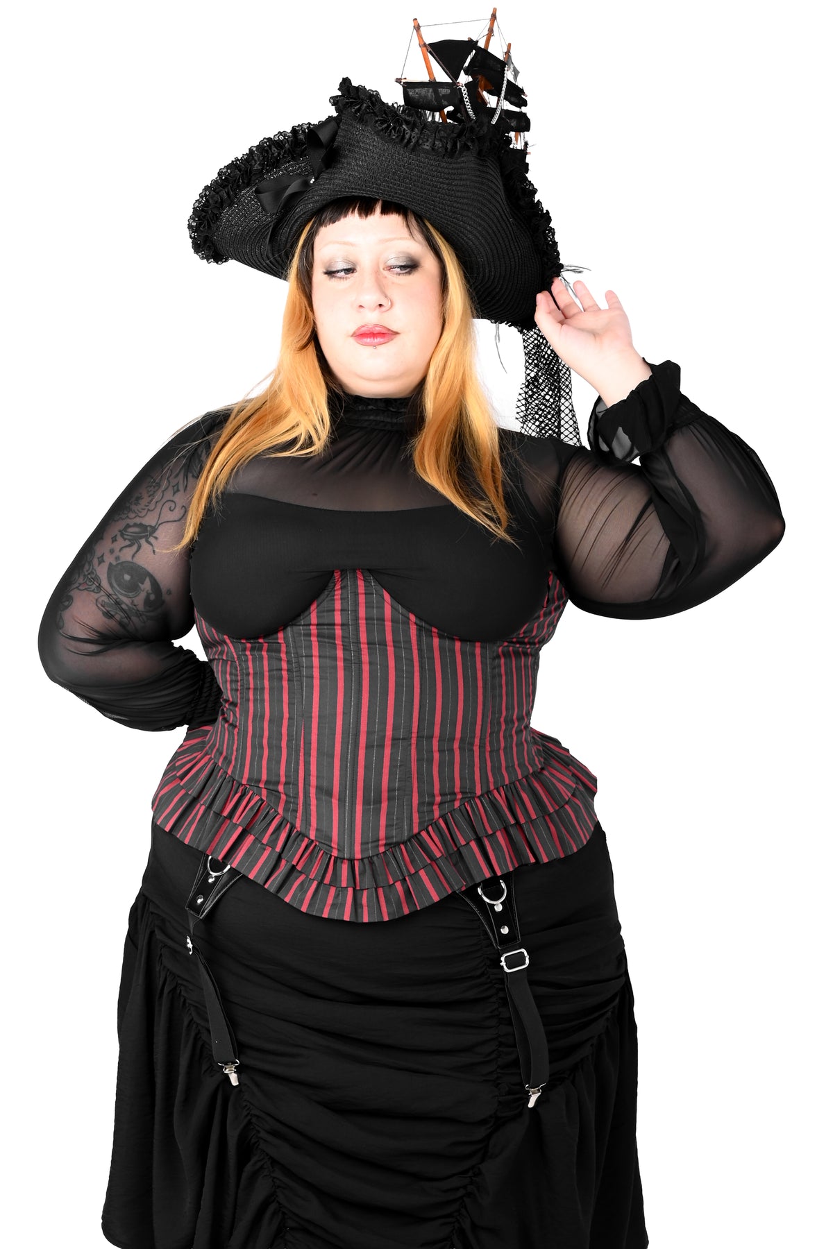 A woman modeling a black pirate costume with a striped corset top, sheer black sleeves, a black skirt, and a black pirate hat with a small ship decoration, against a plain white background.